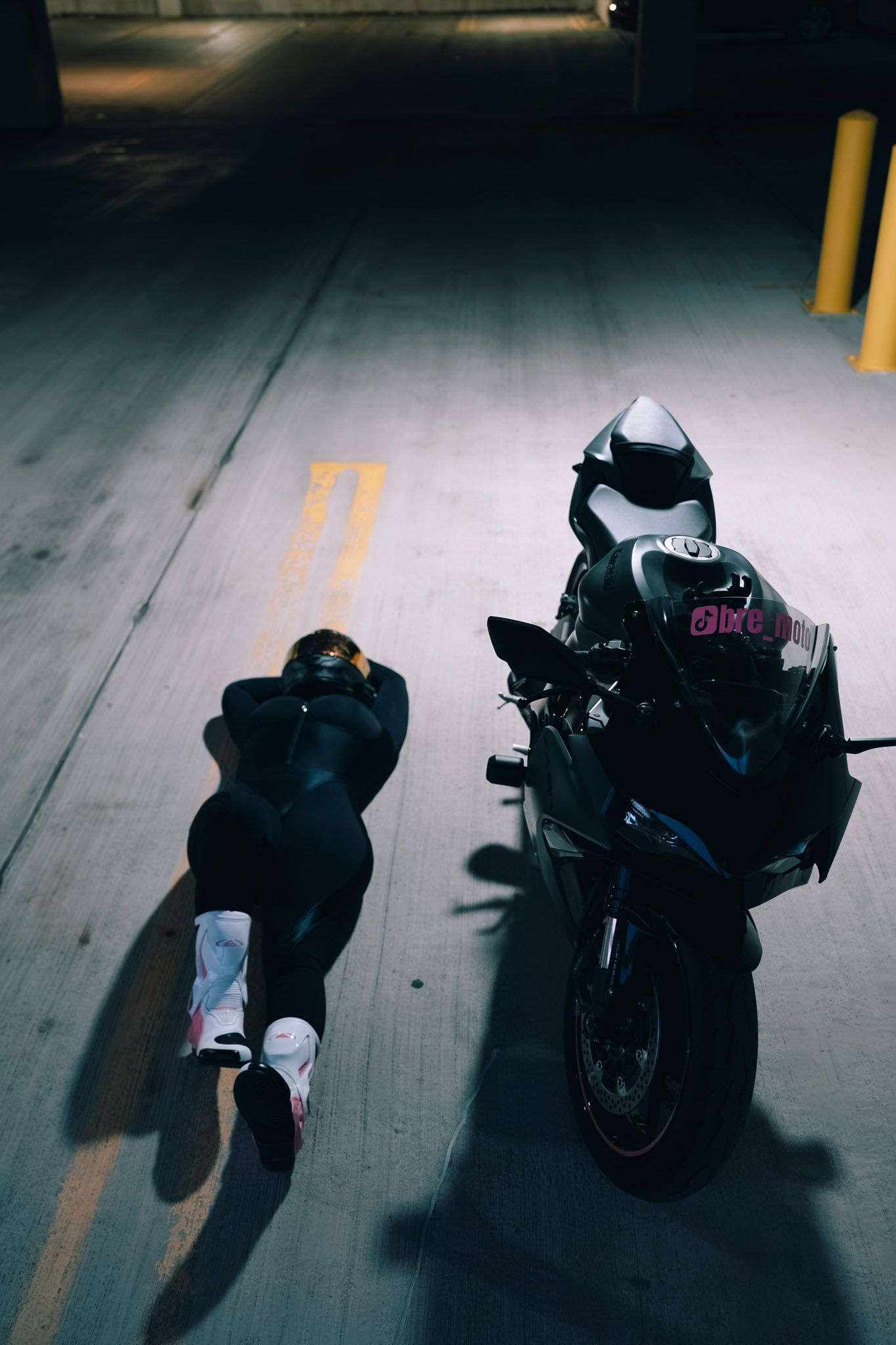 Person in black outfit lying on parking garage floor next to a sport motorcycle at night.