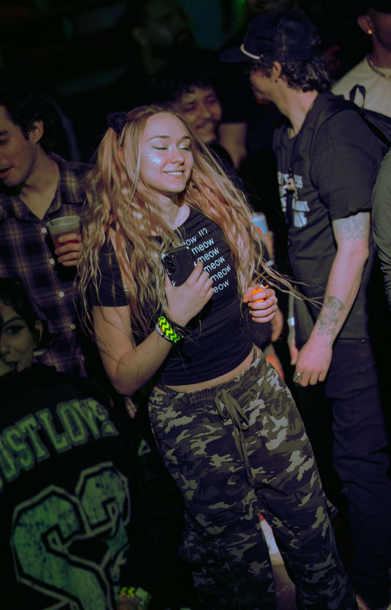 Young woman with long wavy hair dancing at a nightclub, wearing camo pants and a black crop top.