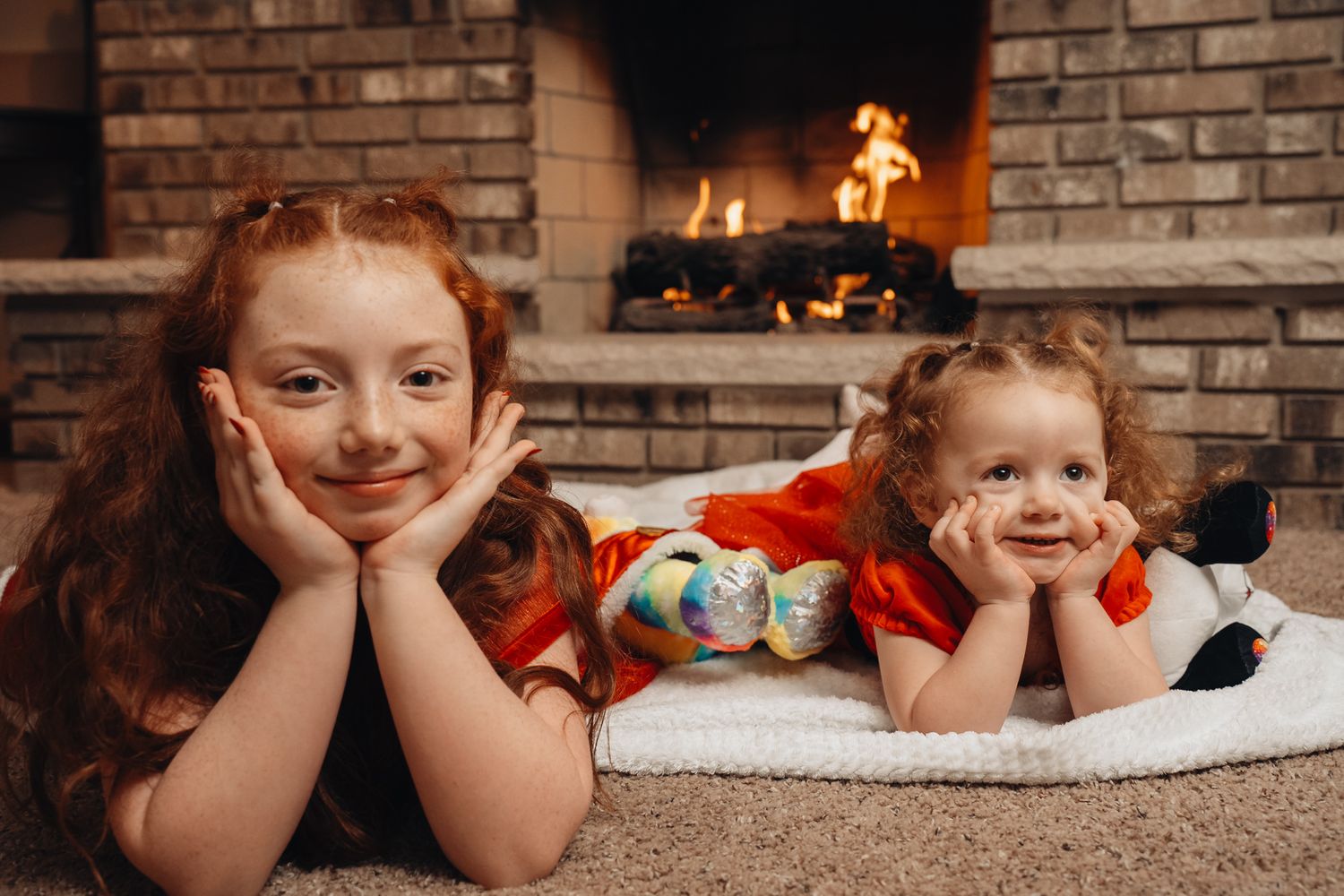 Two children lying on a white rug in front of a brick fireplace, both resting their heads in their hands with cheerful expressions.