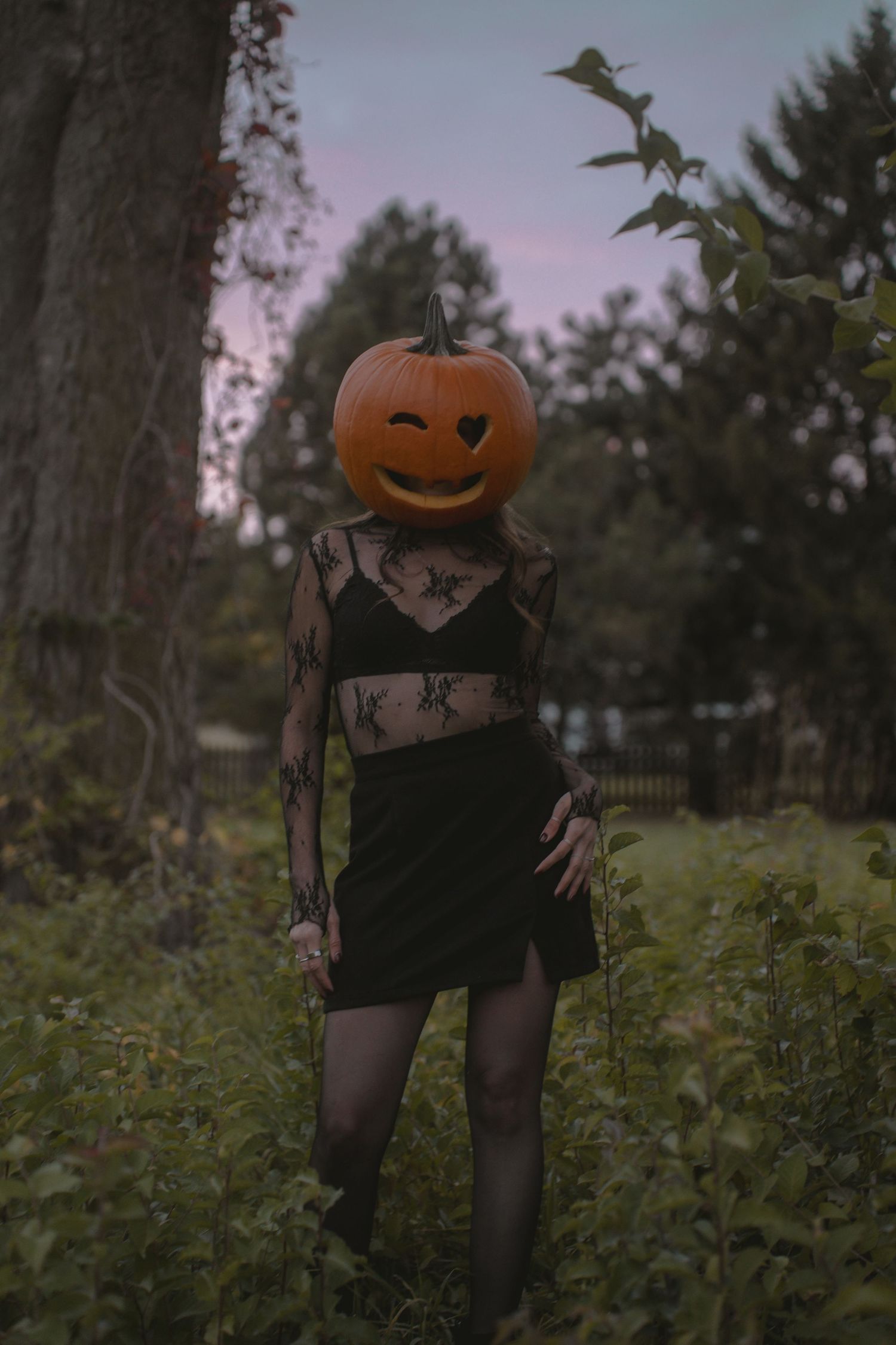 A Halloween-themed figure wearing black clothing and a jack-o'-lantern head poses outdoors in a natural setting.