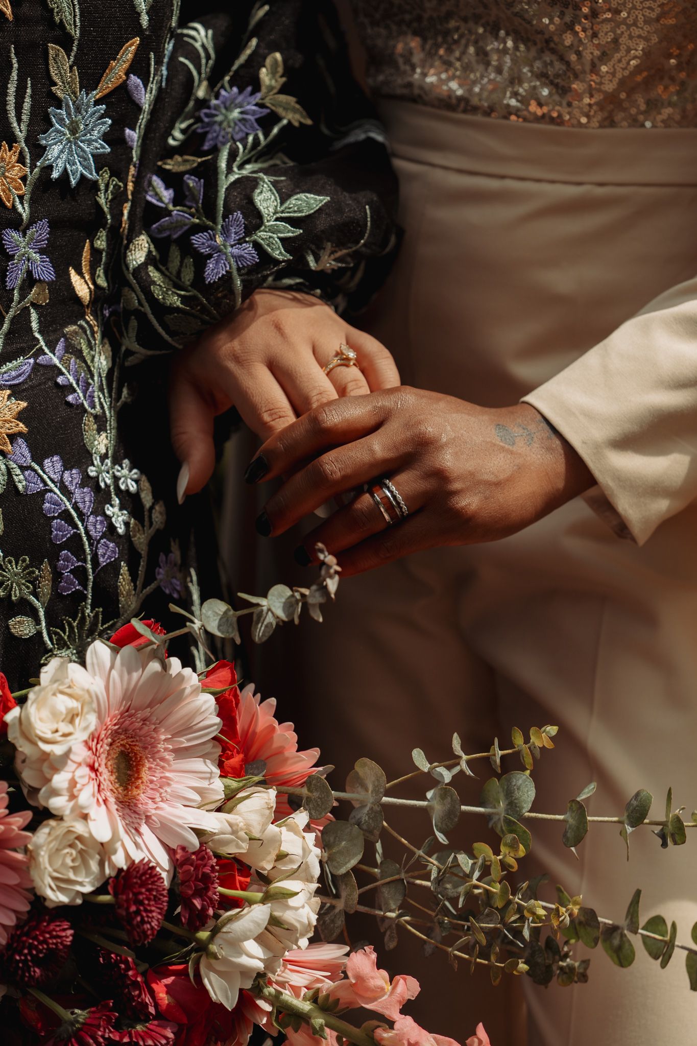 Close-up of couple holding hands with wedding rings, beside a vibrant bouquet of pink and red flowers.
