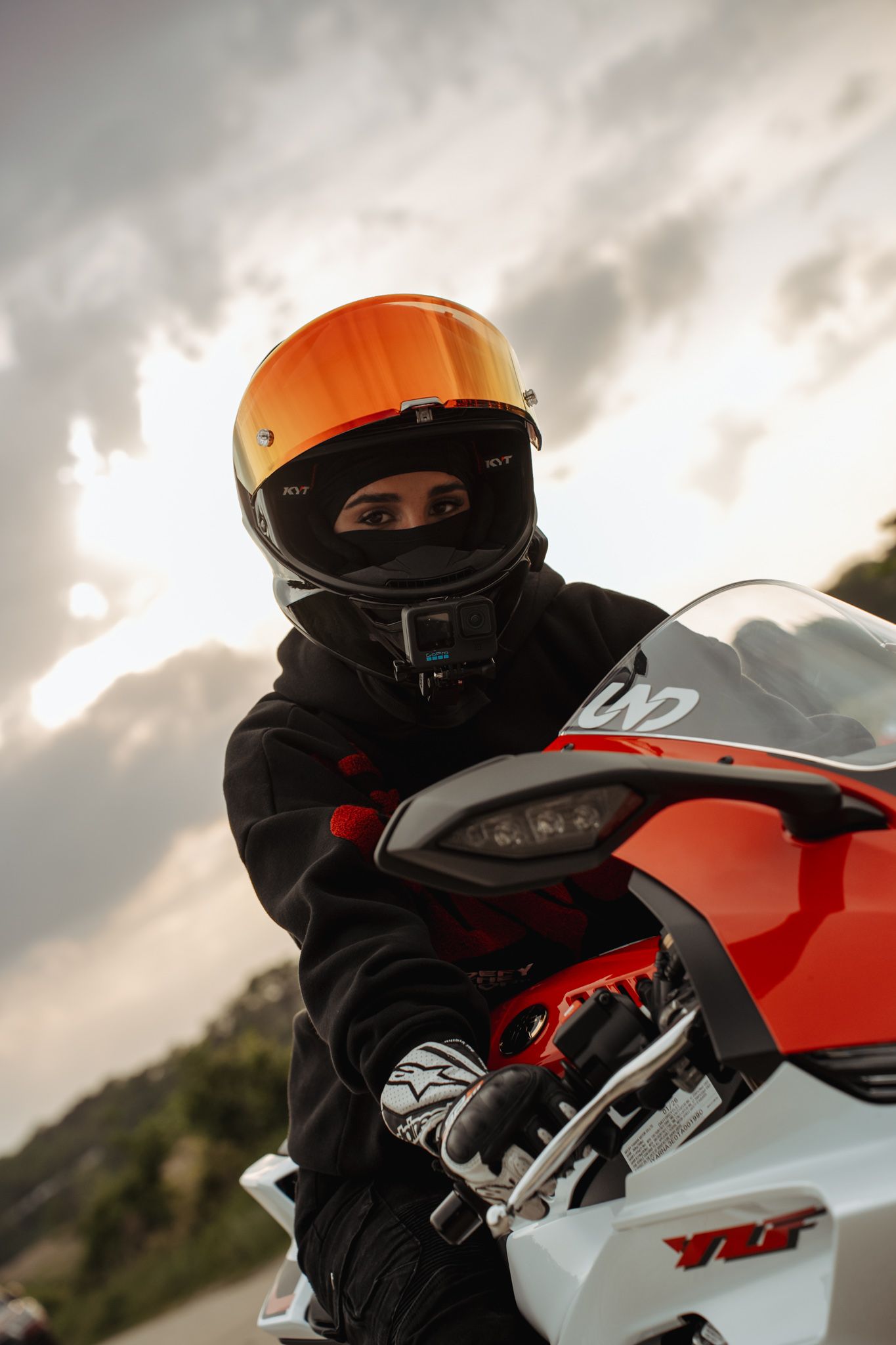 Motorcyclist wearing an orange helmet and black gear poses on a red and white YZF sport bike under cloudy skies.