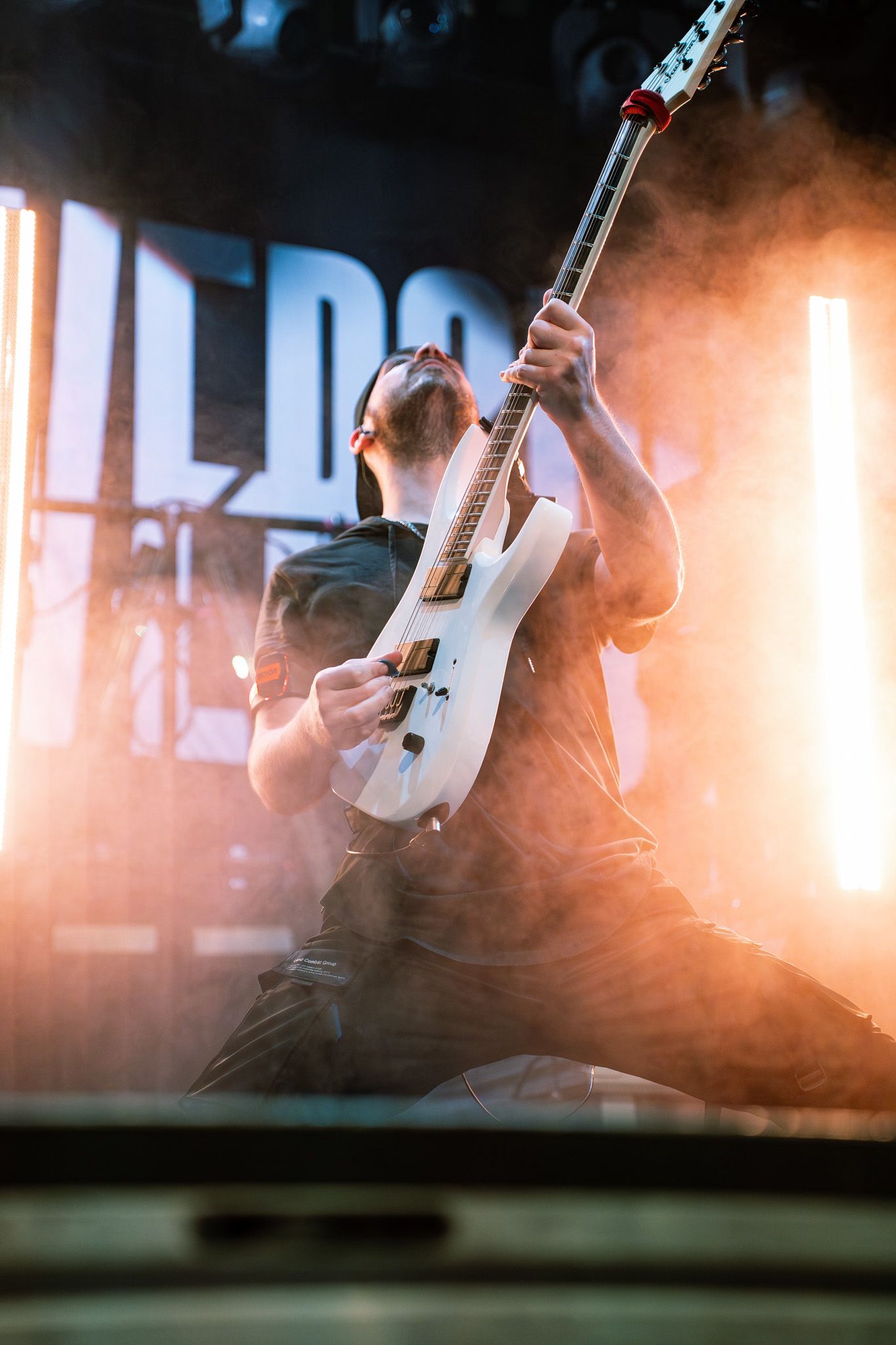 Guitarist performs energetically on stage with a white electric guitar backlit by dramatic orange lighting and smoke effects.