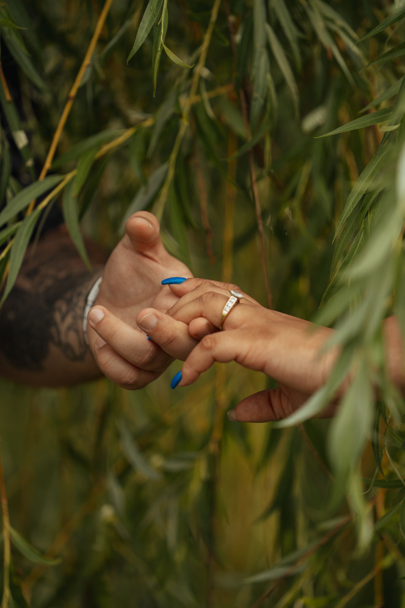 Tattooed man placing a gold ring on a woman's finger with blue nails, surrounded by green willow leaves.