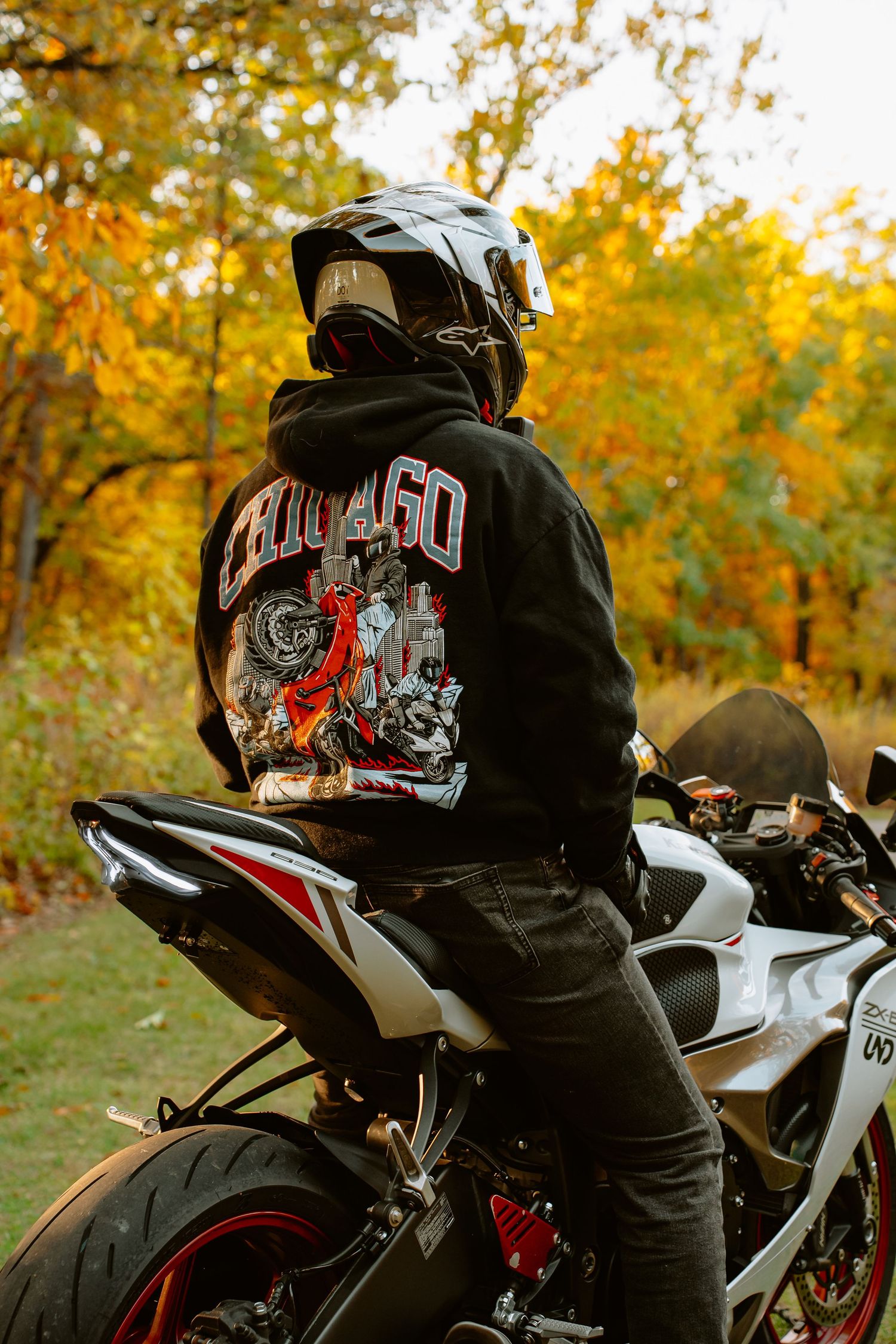 A motorcyclist in a black hoodie rides a white sportbike against a backdrop of autumn trees with golden foliage.
