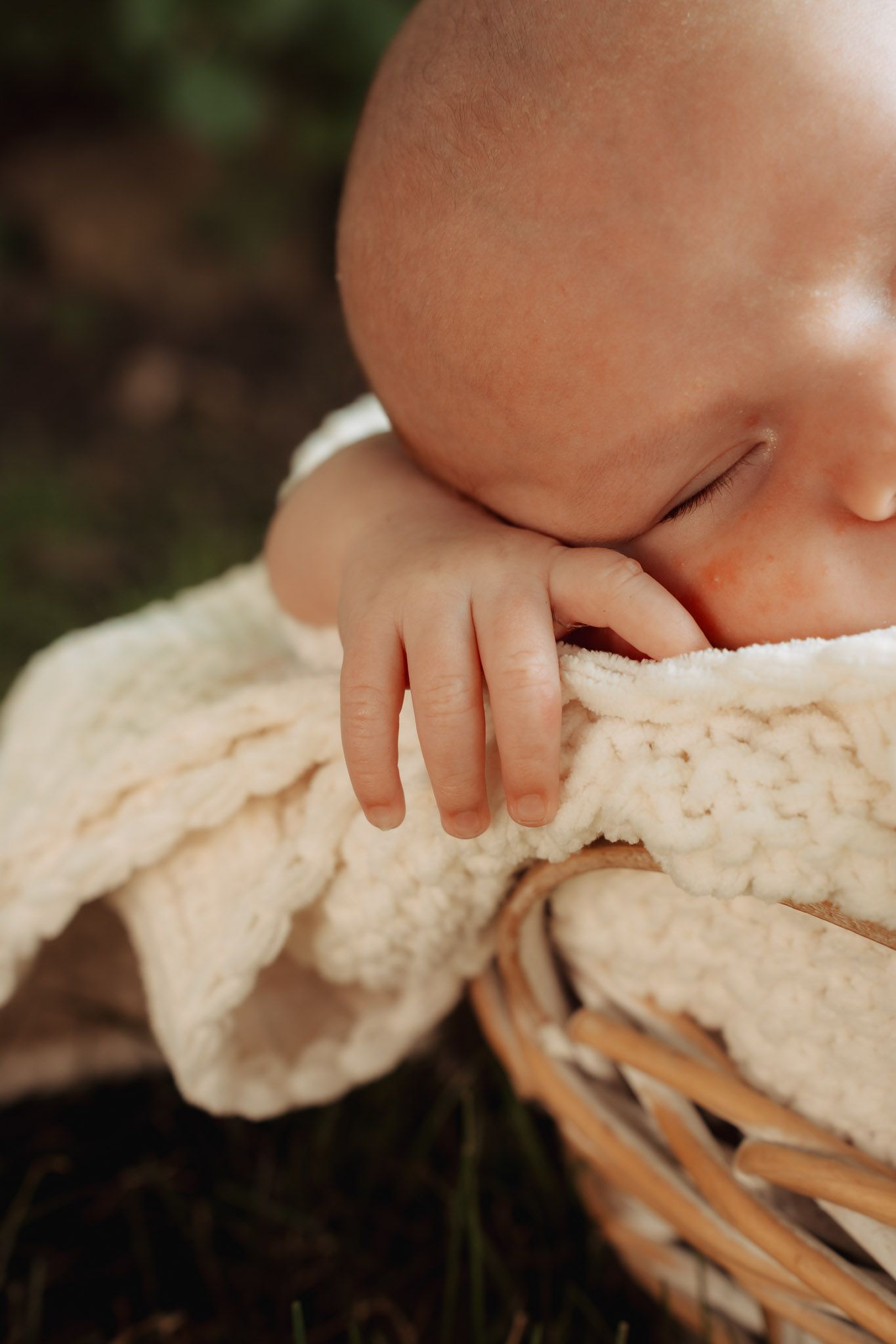 Close-up of a sleeping newborn baby resting chin on hands over a cream knit blanket in a wicker basket outdoors.