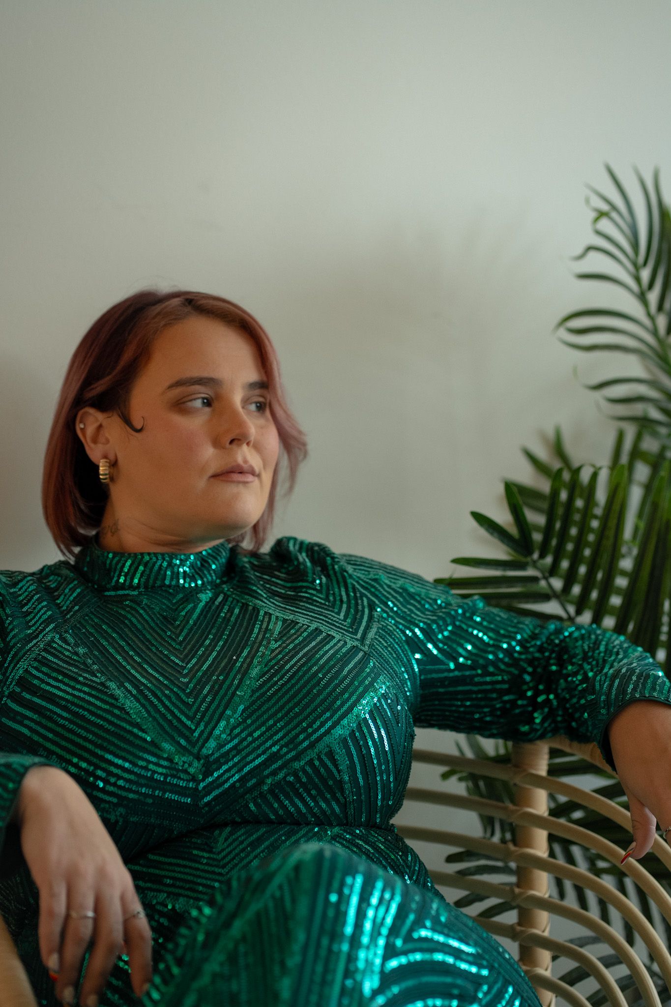A person in a sparkling emerald green sequined dress poses against a white wall with tropical plants.