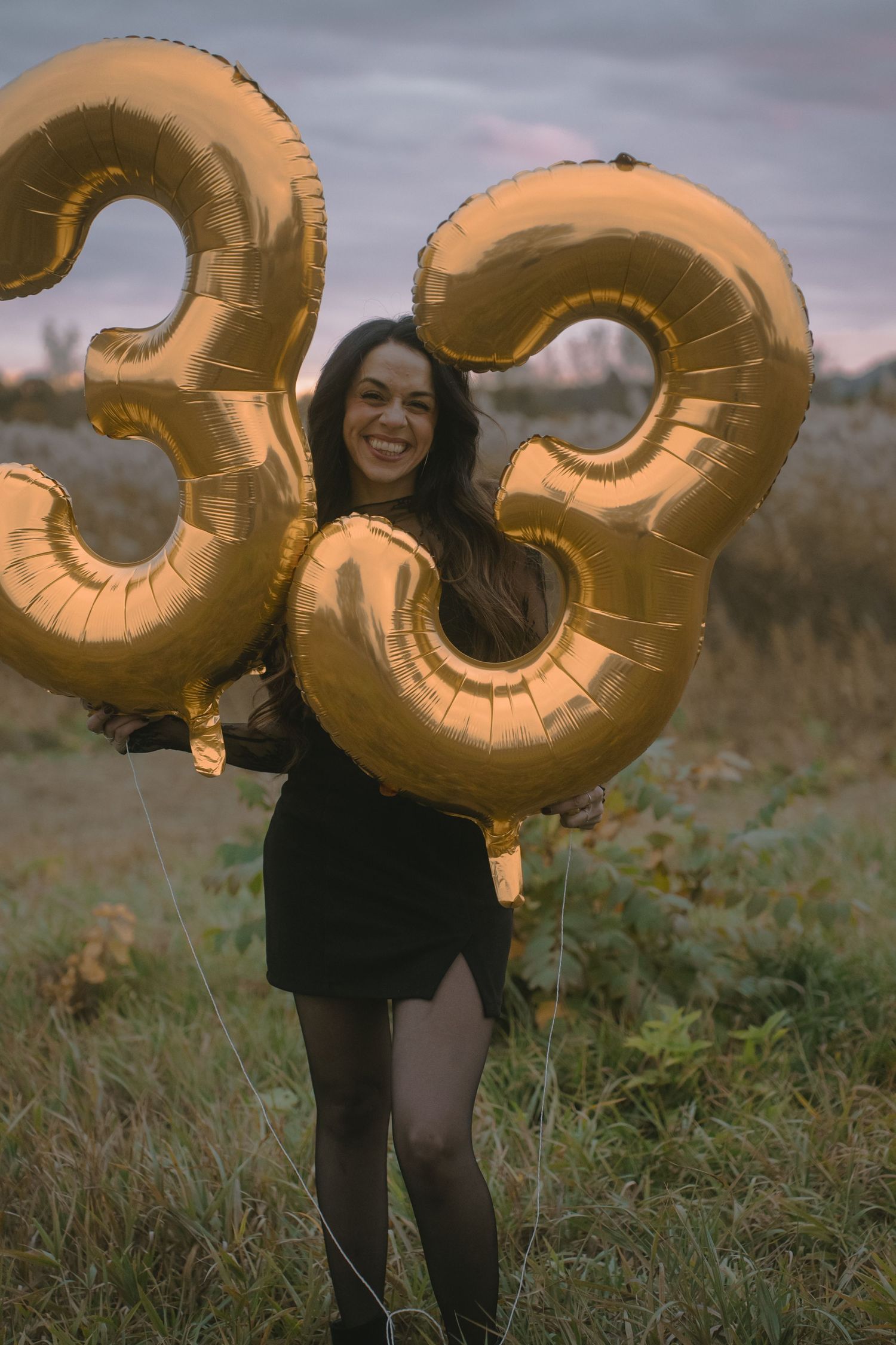 A woman in black holds large gold number 33 balloons in an autumn field at sunset.