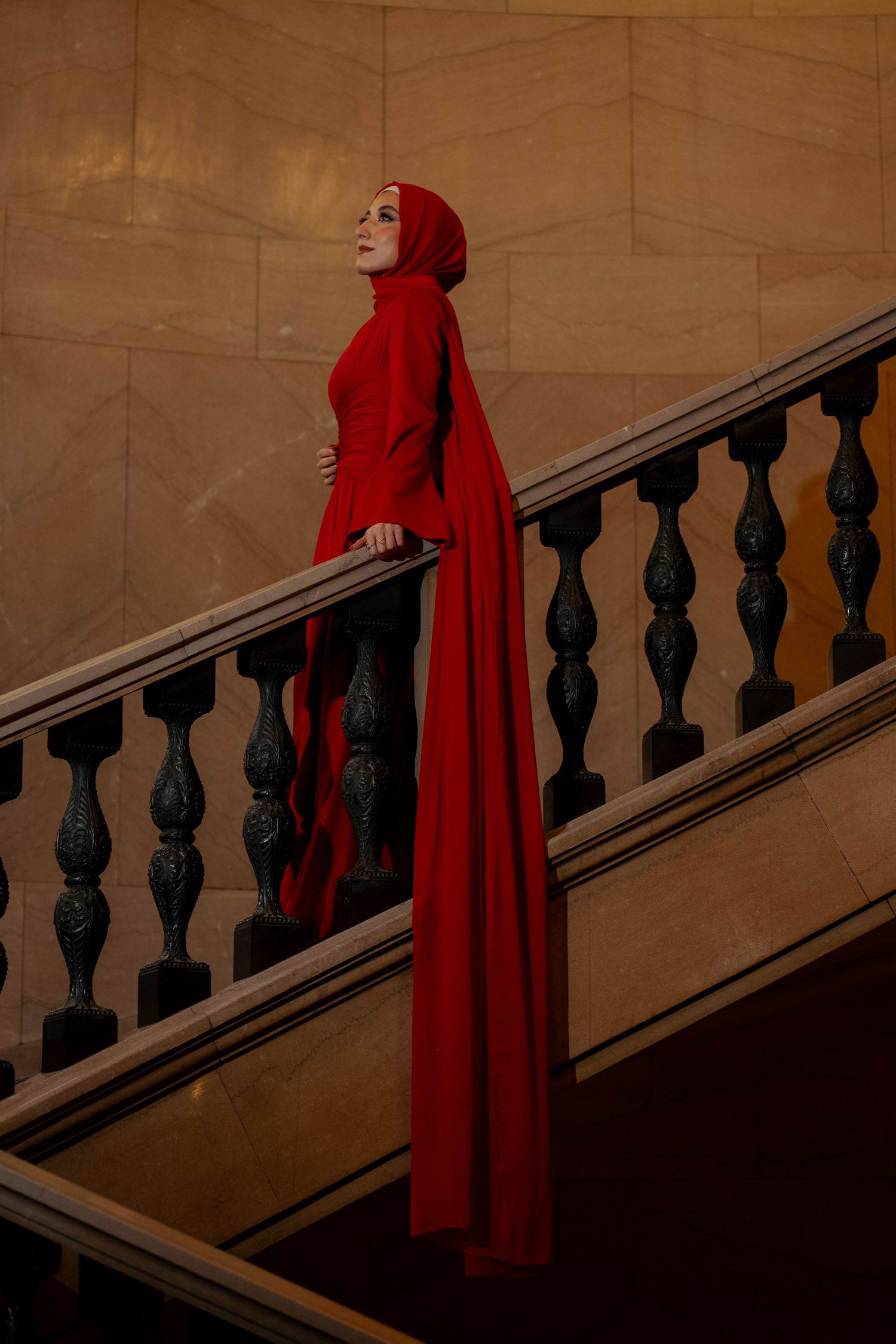 Woman in elegant red hijab and flowing gown poses gracefully on ornate marble staircase.