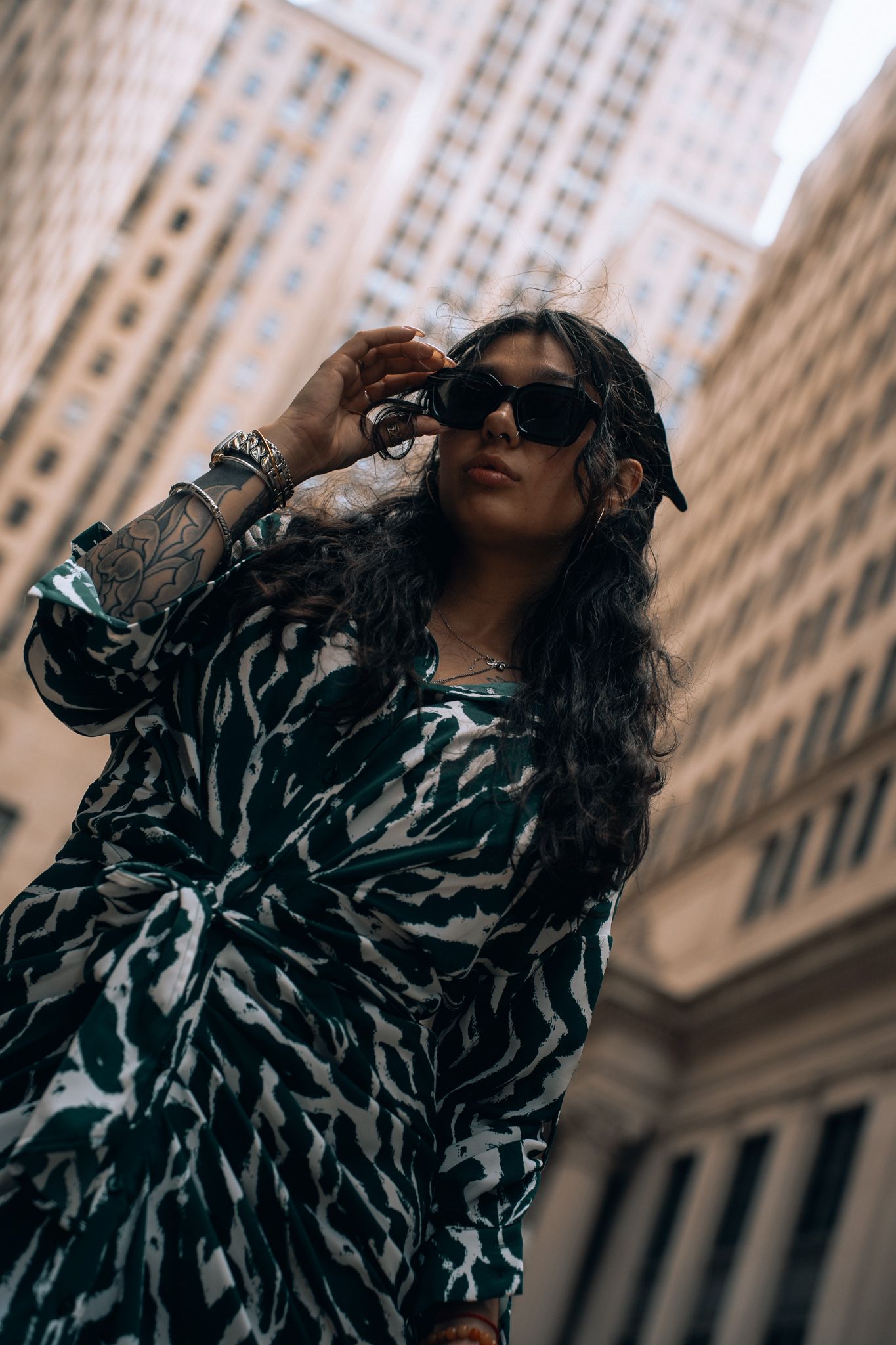 Tattooed woman in zebra print dress and black cap adjusts sunglasses against urban skyscraper backdrop.