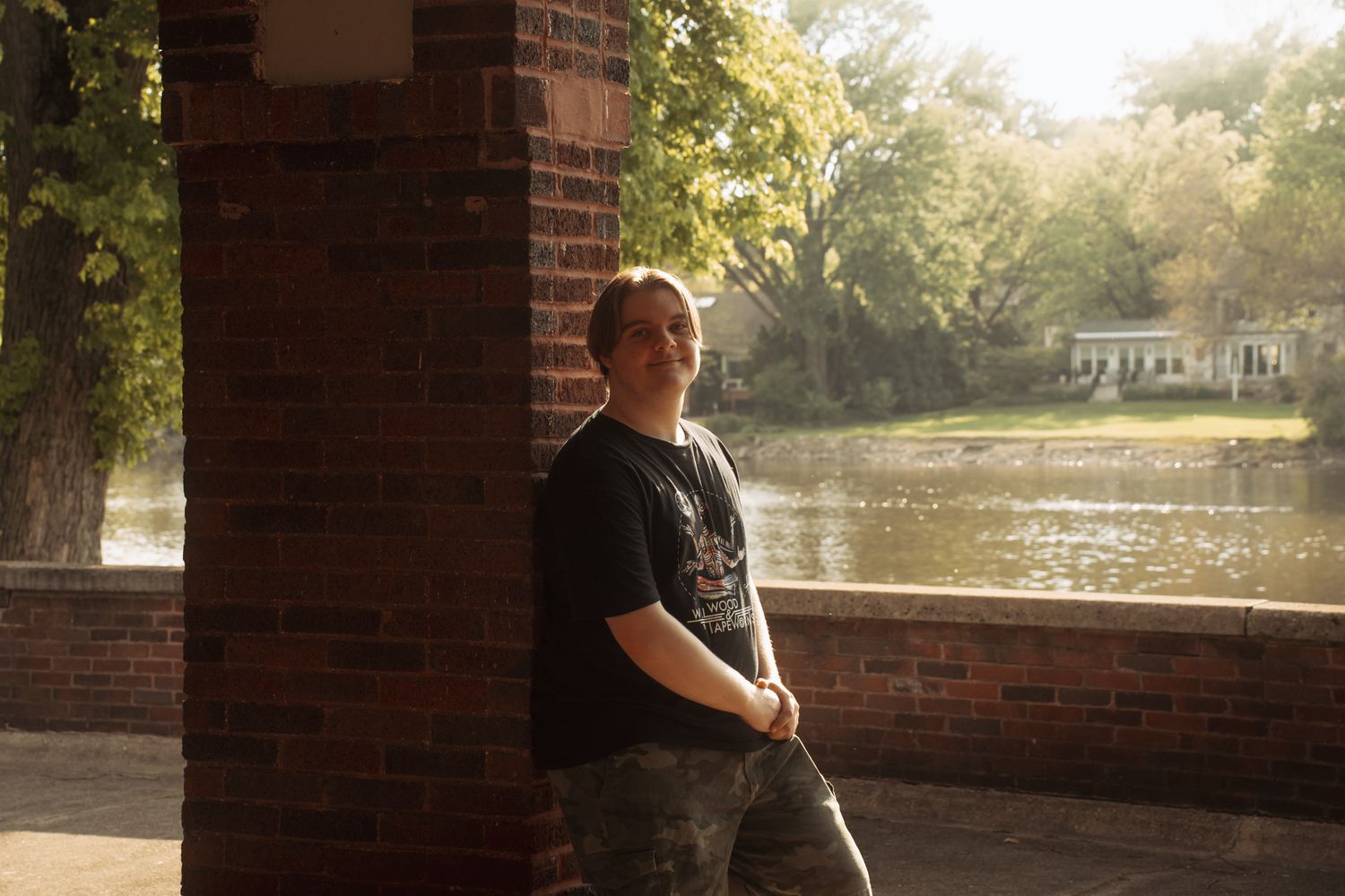 Man in black t-shirt leaning against brick column near scenic river with trees and white building in background.