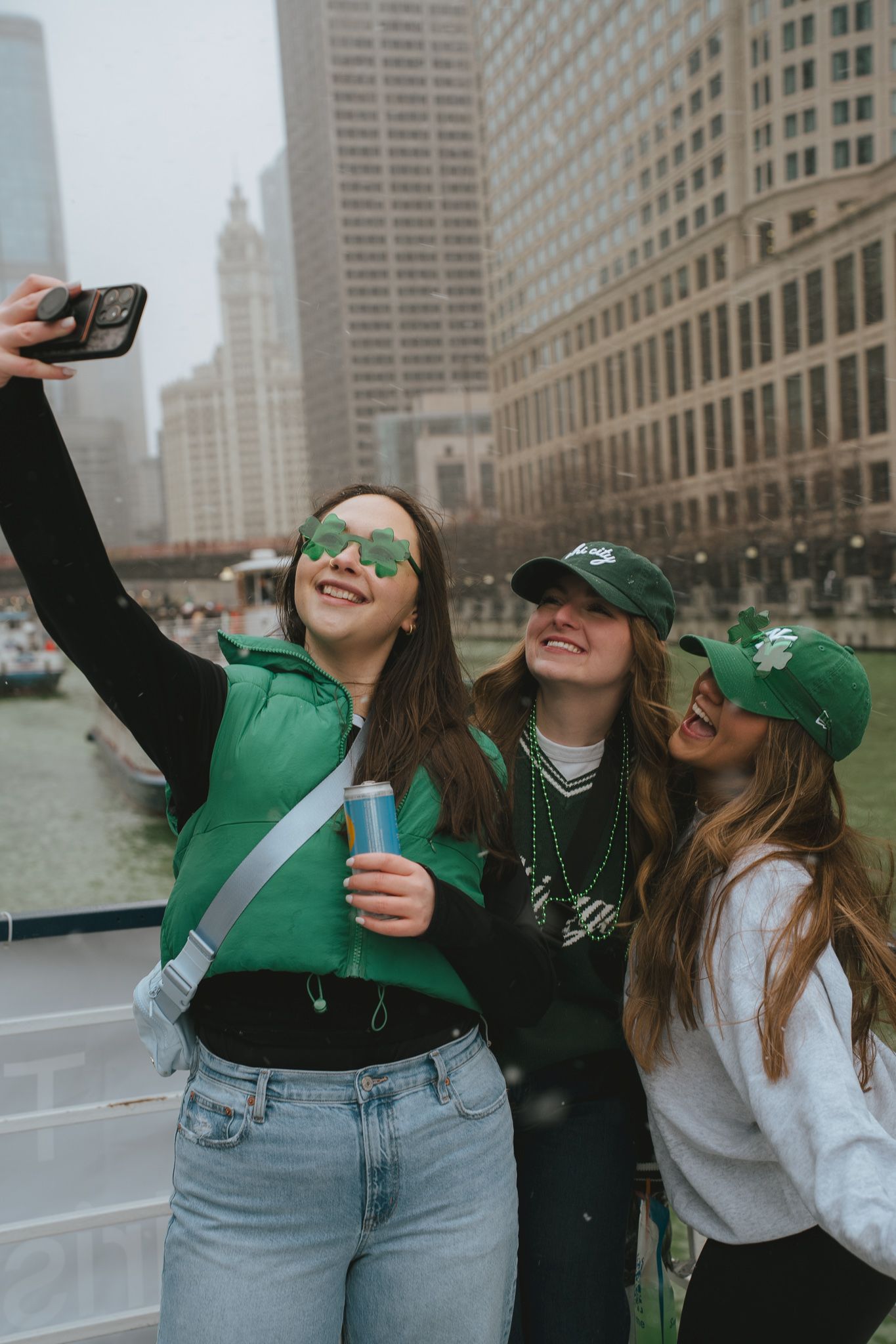 Friends in green St Patrick's Day attire taking selfies along Chicago River with downtown buildings in background.