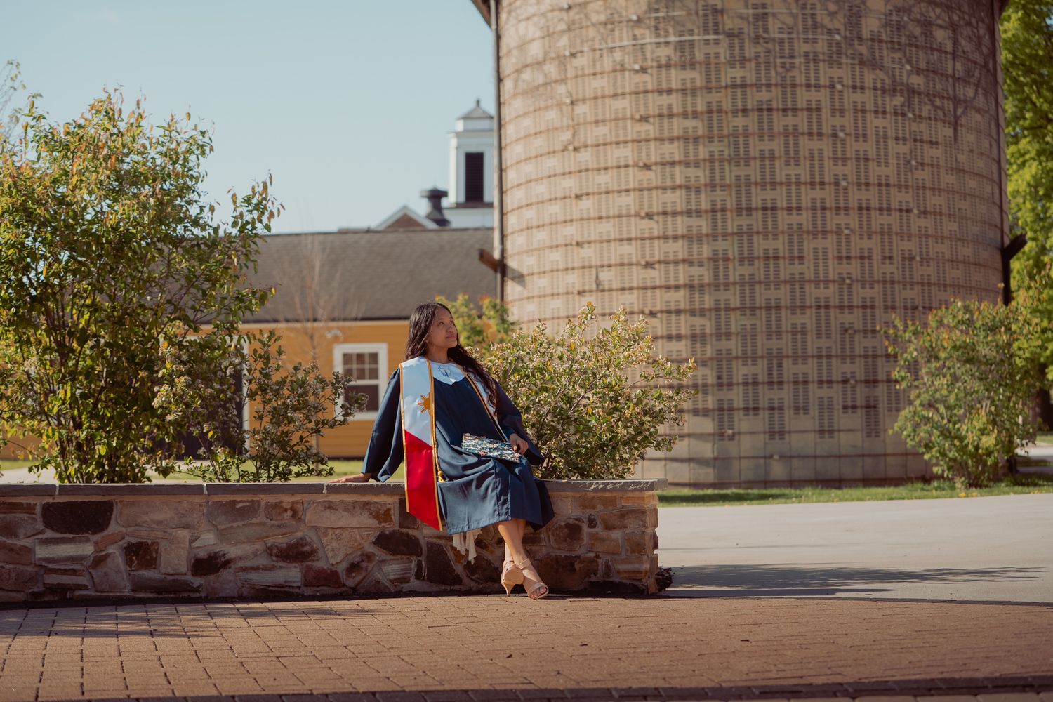 Woman in graduation gown walks past a stone wall and silo on a sunny college campus.