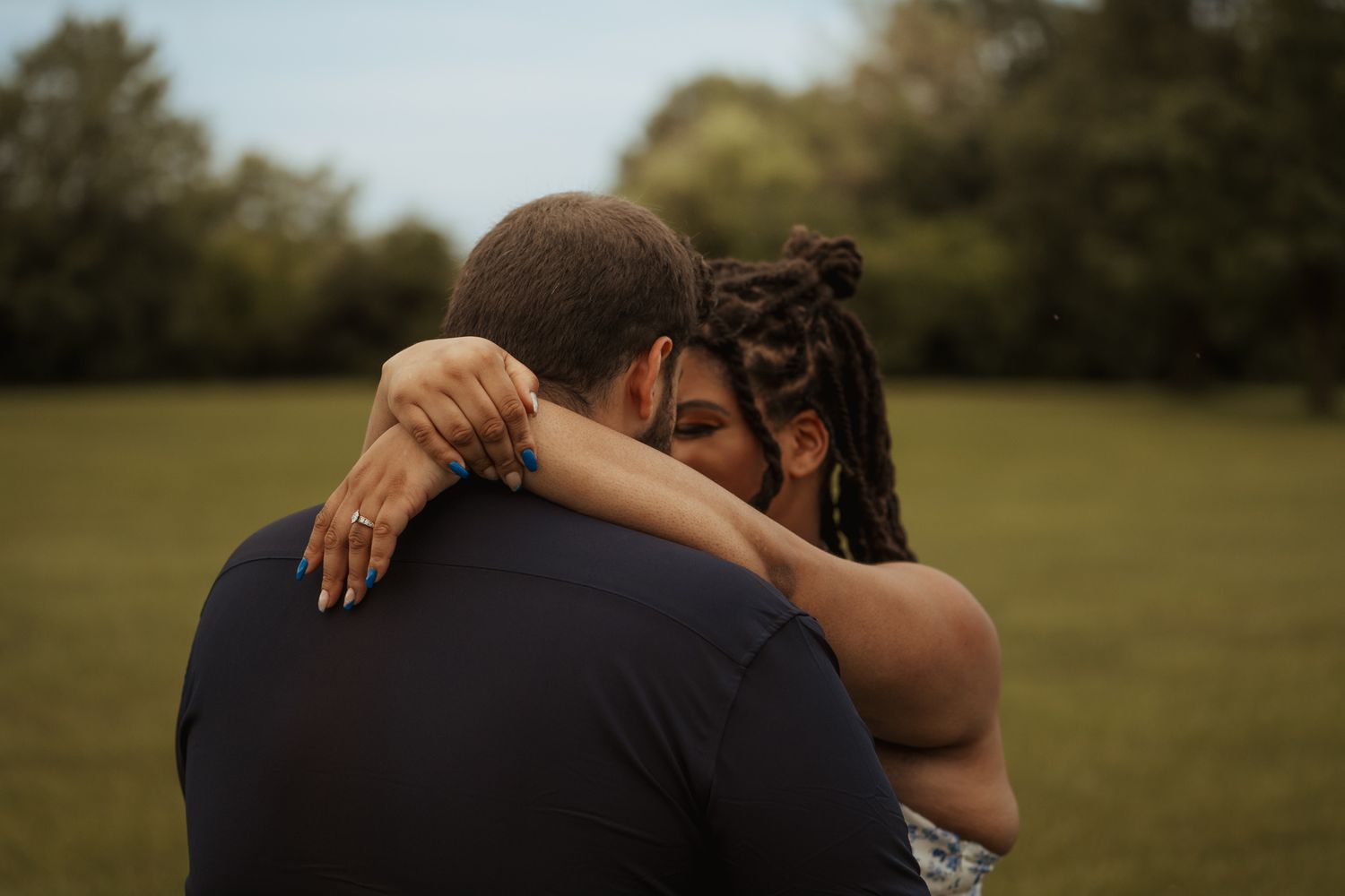 A couple shares a tender embrace outdoors in a green field, woman with locs wrapping arms around man in navy shirt.