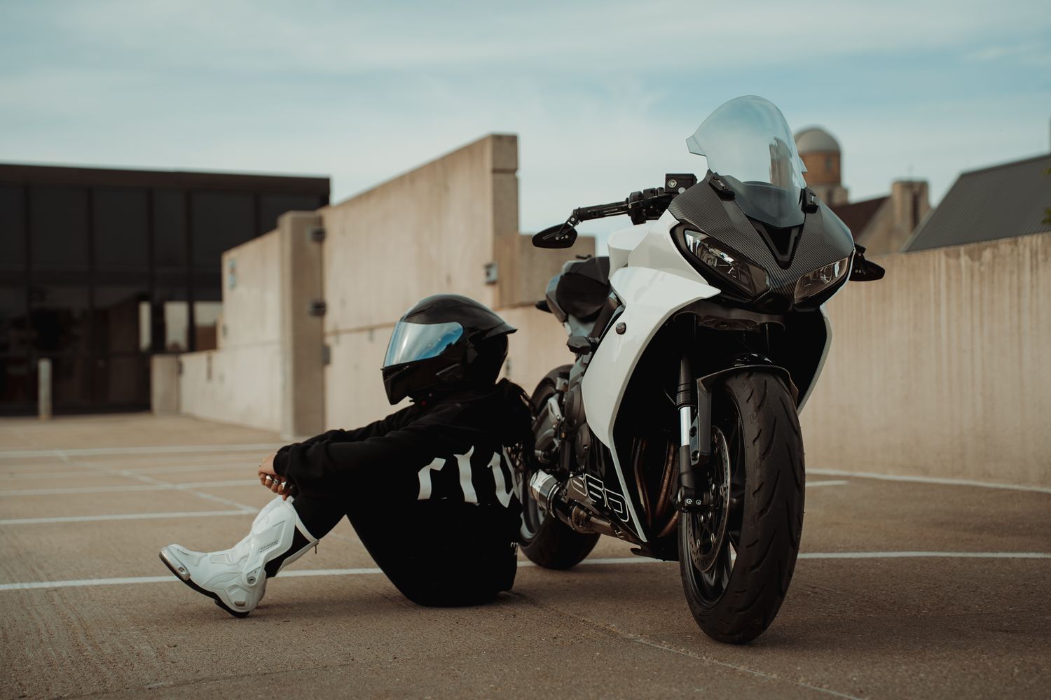 A motorcyclist in black gear and helmet sits beside a white sport motorcycle on a rooftop parking lot.