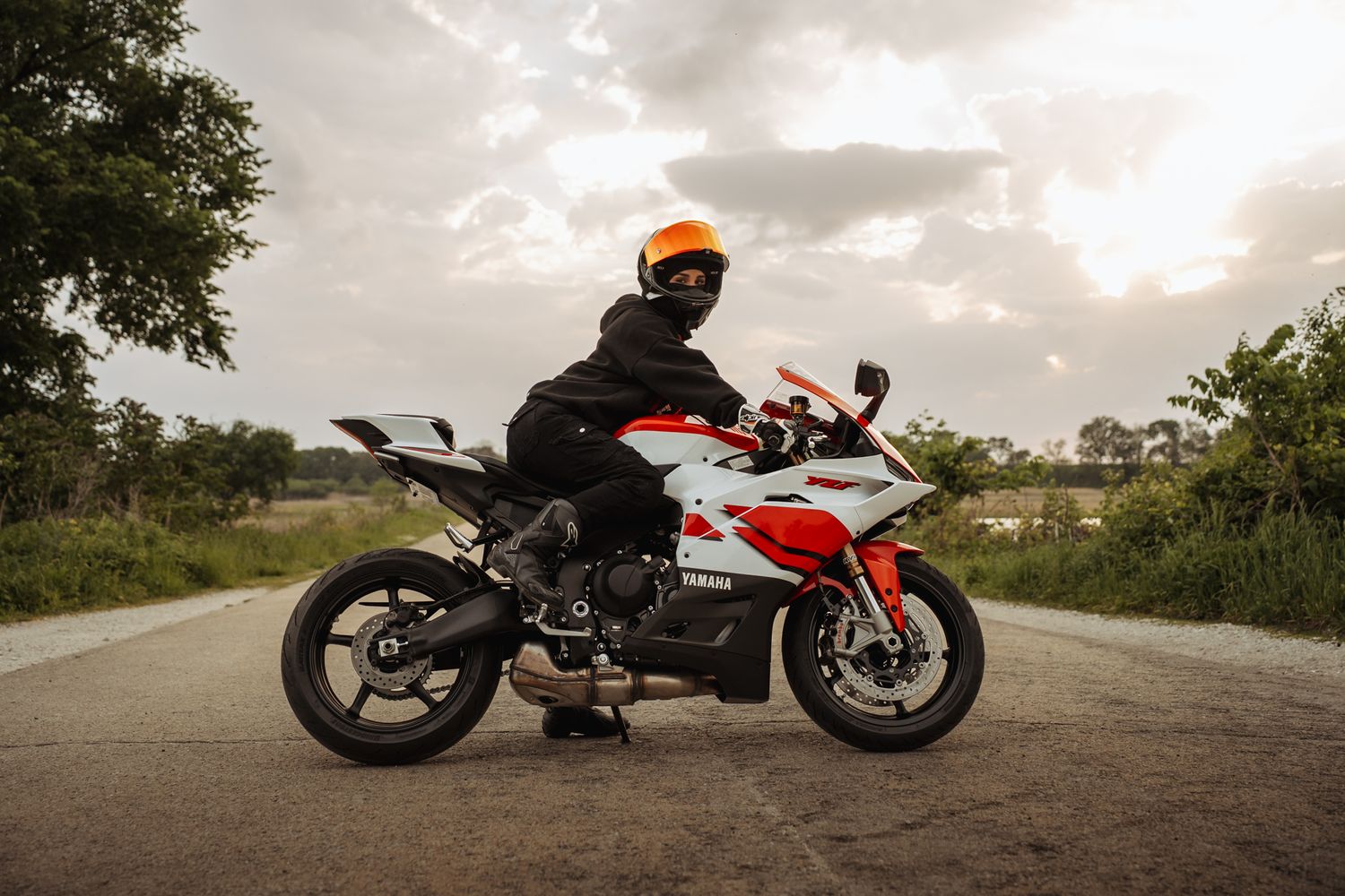 Motorcyclist in orange helmet and black jacket sitting on a white and red Yamaha sport bike on a rural road.
