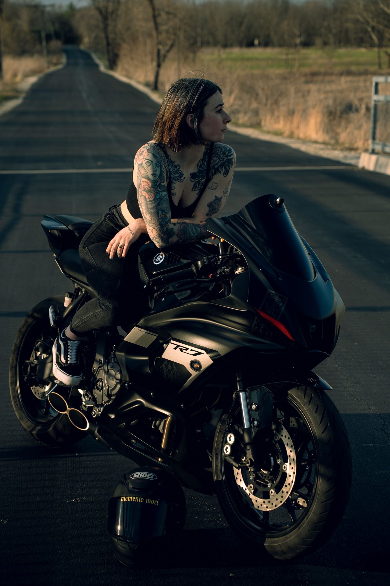 Tattooed woman leaning on a black Yamaha R6 sport motorcycle parked on an empty road at dusk.