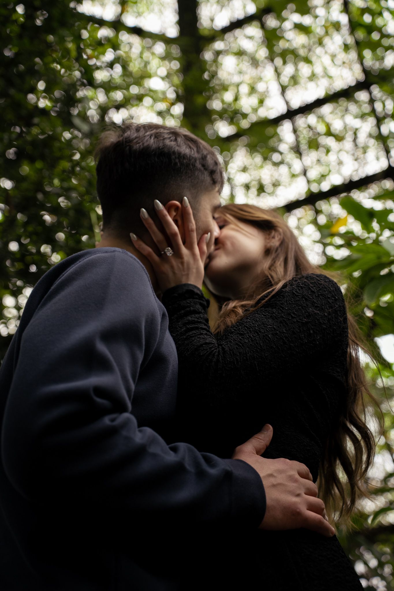 Two silhouettes embrace in a romantic kiss against a sunlit backdrop of green foliage.