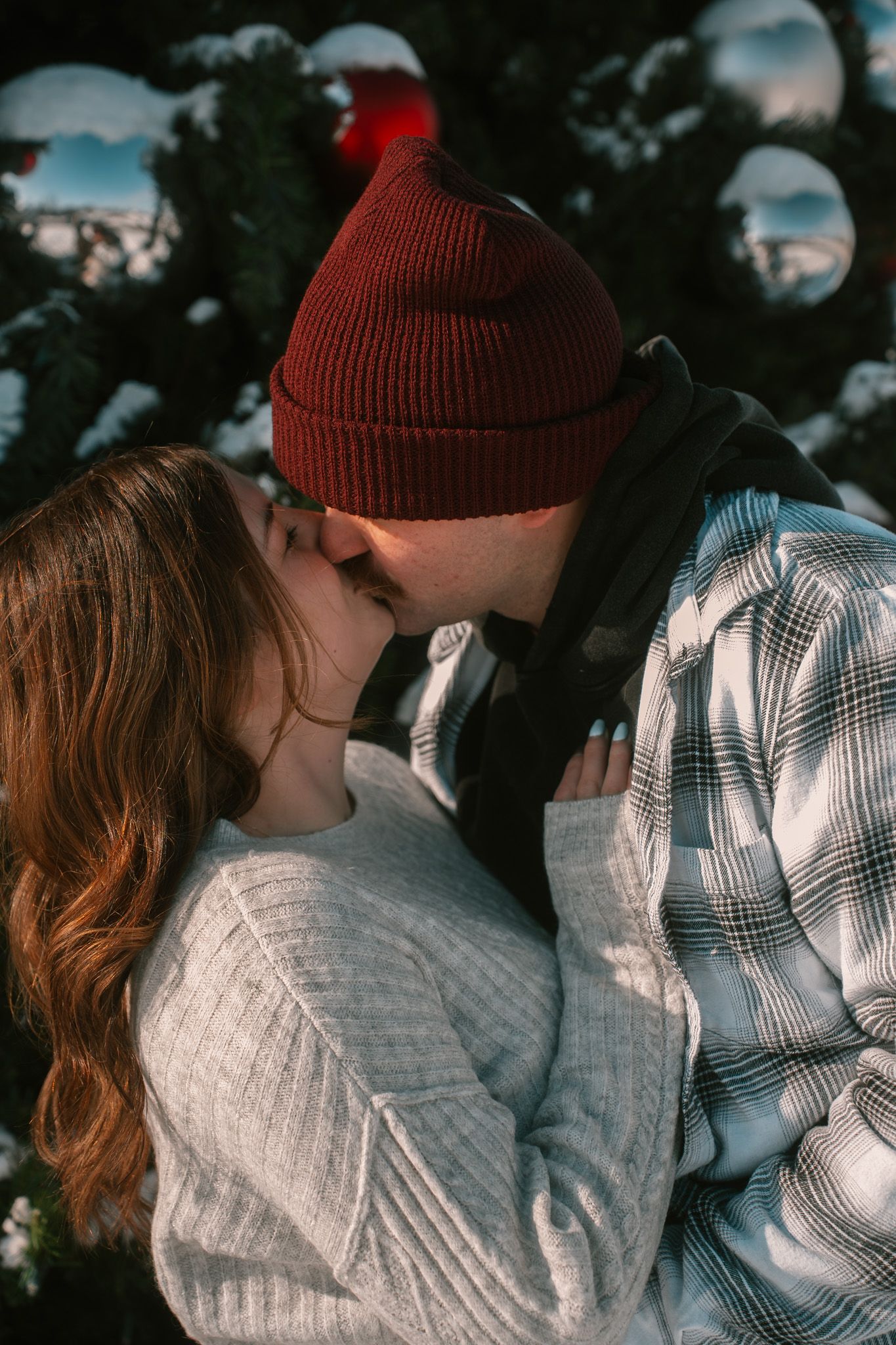 A couple shares a romantic winter kiss outdoors, wearing cozy sweaters and a red beanie against a snowy background.