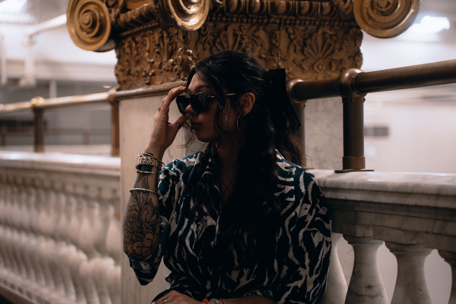 A tattooed woman in a zebra-print blouse and sunglasses leans on a marble balustrade in an ornate interior.