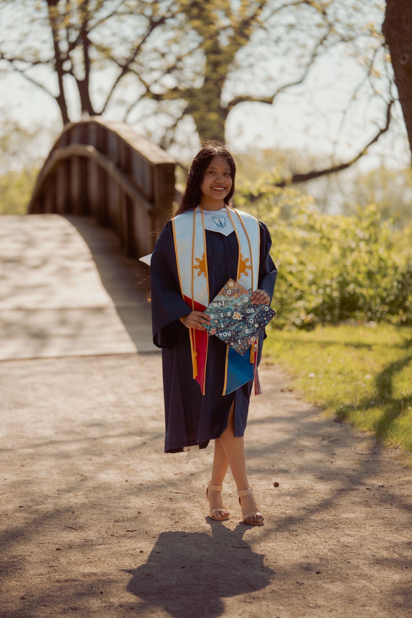 Smiling graduate in navy cap and gown with colorful stole holds decorated diploma outdoors near wooden bridge.
