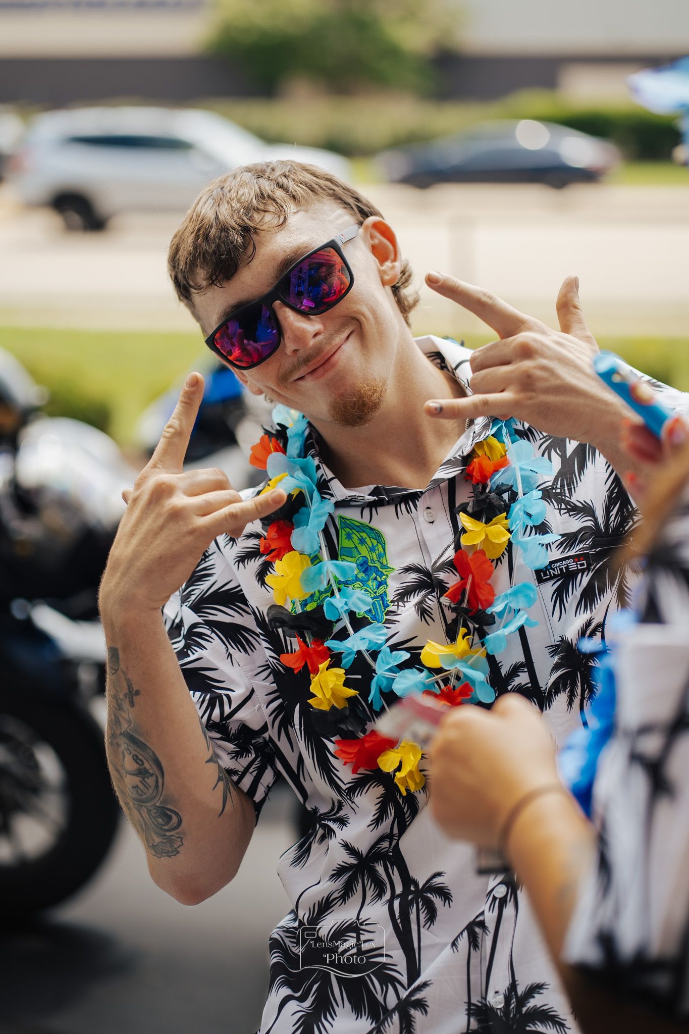 Person in a tropical print shirt and sunglasses wearing colorful leis gestures playfully at an outdoor event.