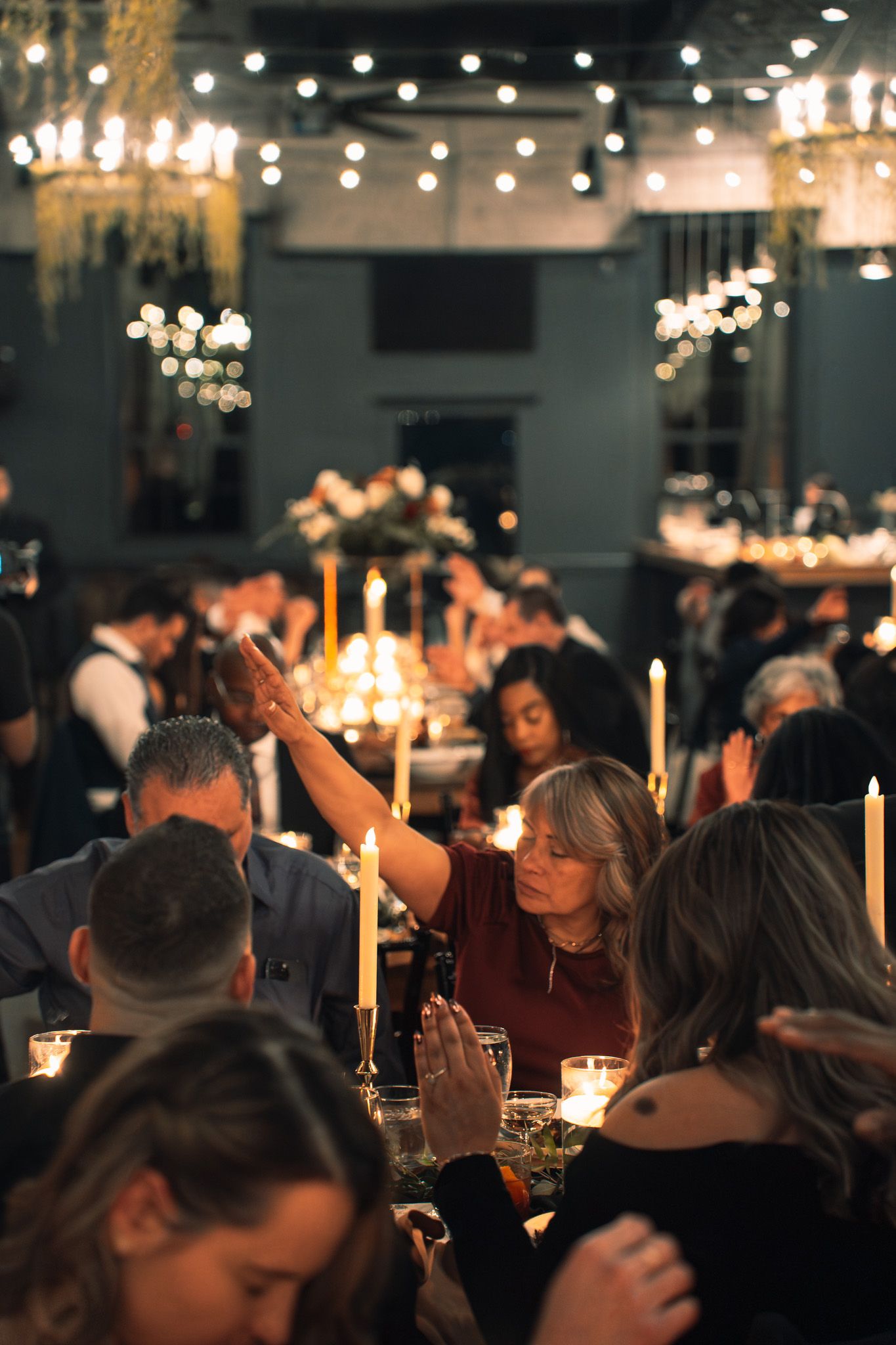 Candlelit dinner party scene with guests seated at tables under warm lighting and string lights creating an intimate atmosphere.