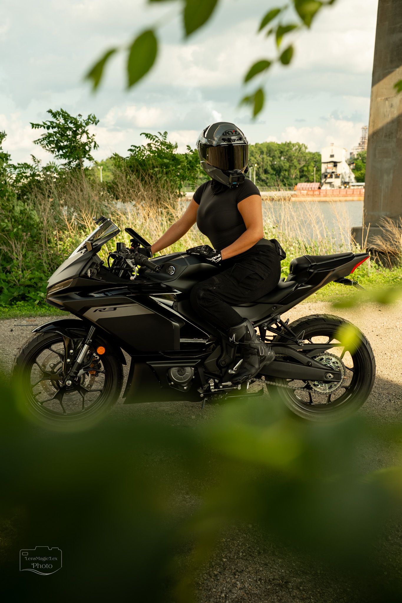 Sport bike rider in black gear crouches low on motorcycle near foliage.