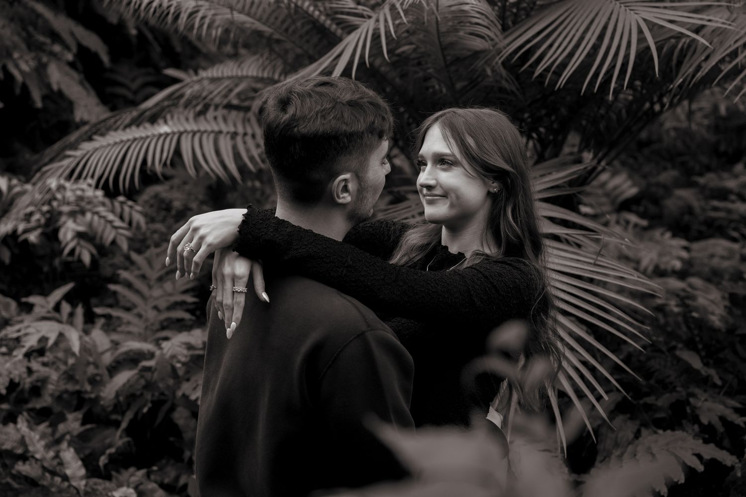 A couple shares an intimate moment together in black and white, surrounded by tropical palm fronds in a garden setting.