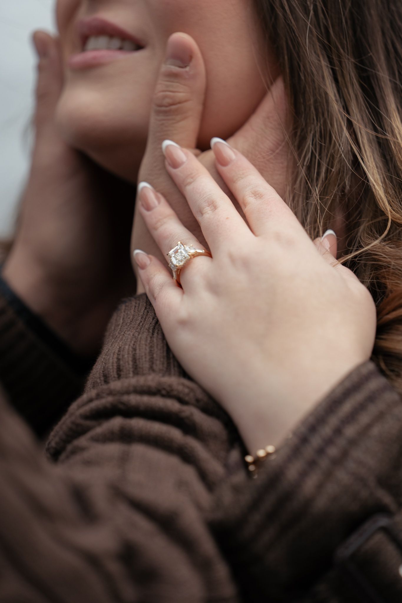 Close-up of hands wearing a diamond engagement ring against a dark brown sweater background.