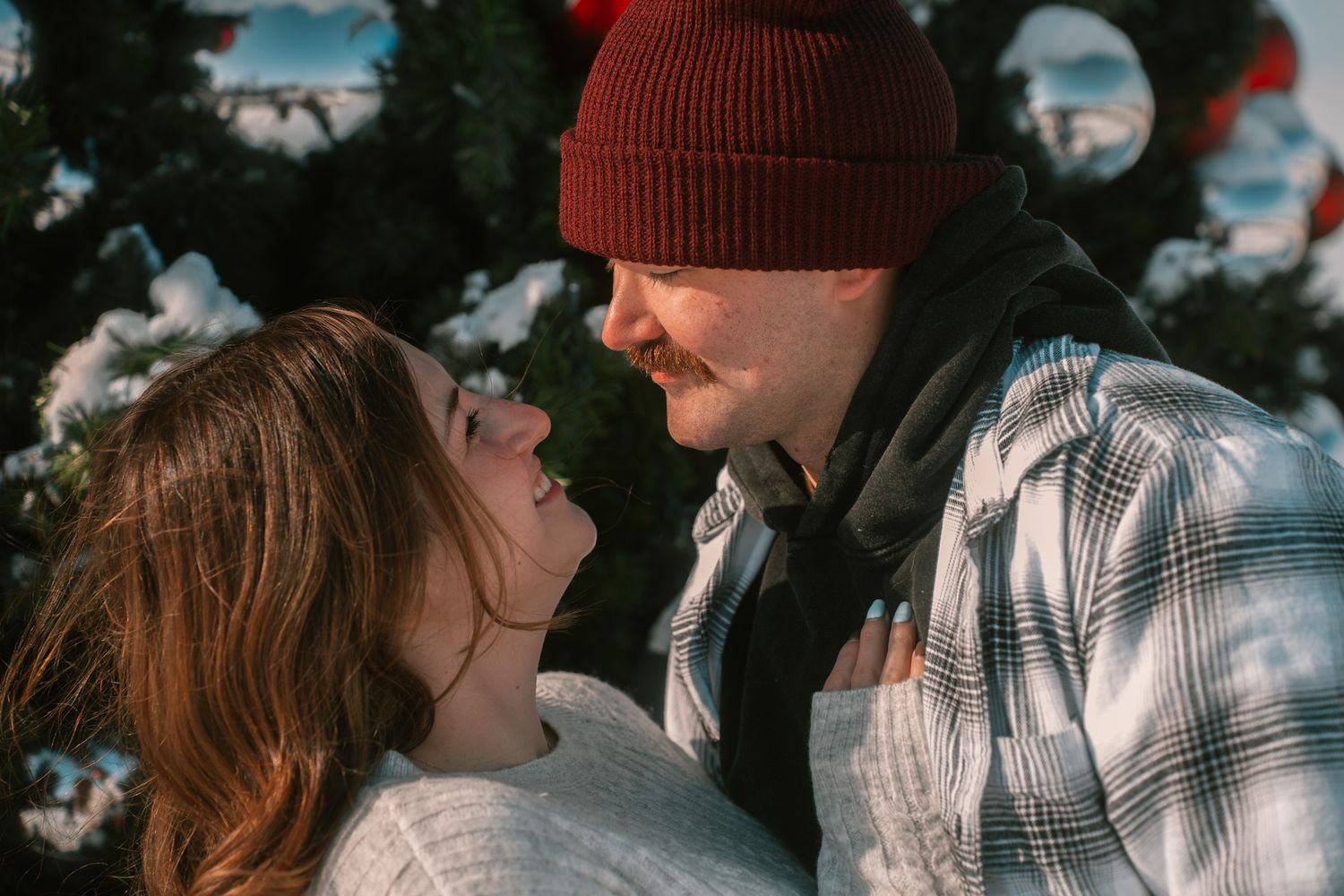 A couple shares a joyful moment outdoors wearing winter attire including a red beanie and plaid jacket.