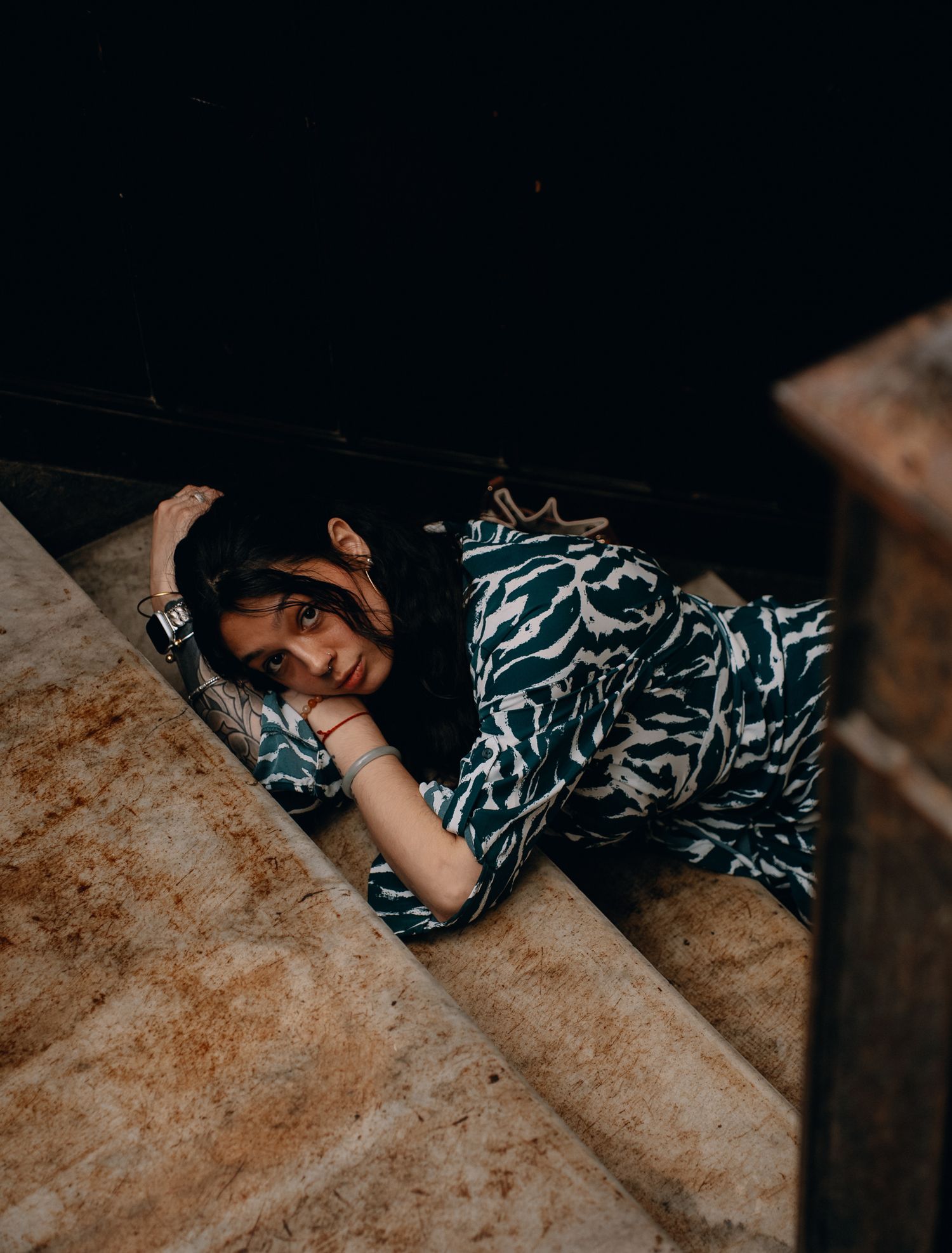 Woman in blue patterned dress lying on stone steps, looking at camera in moody artistic portrait photography.