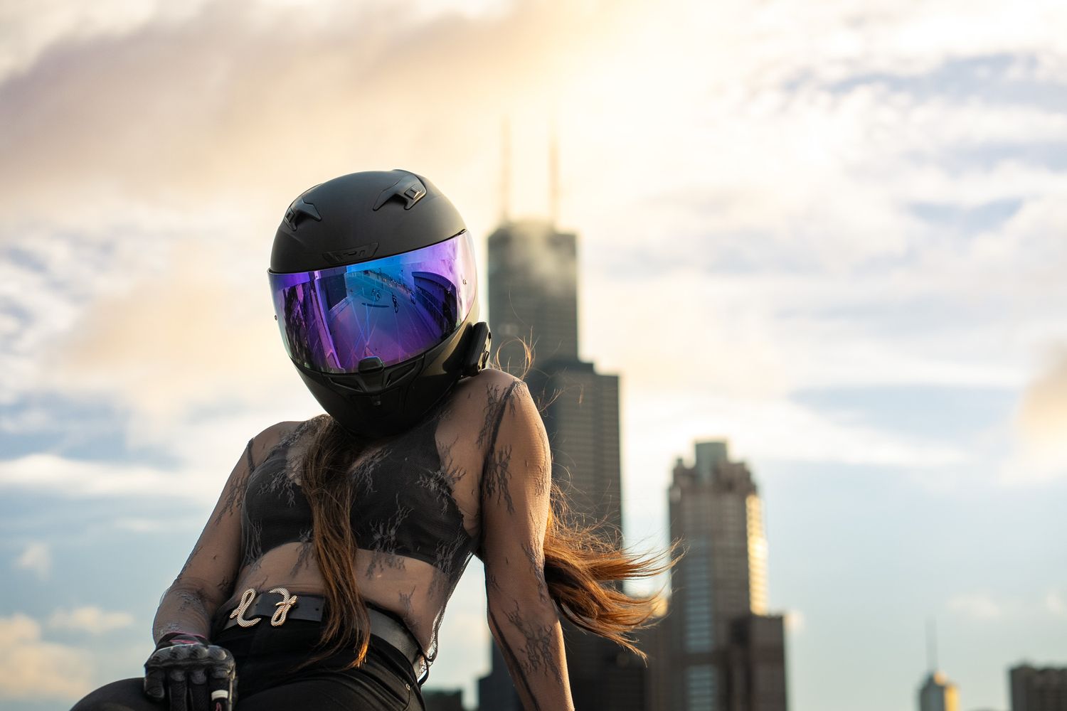 Motorcycle rider in dark gear photographed against warm sunset sky and buildings.