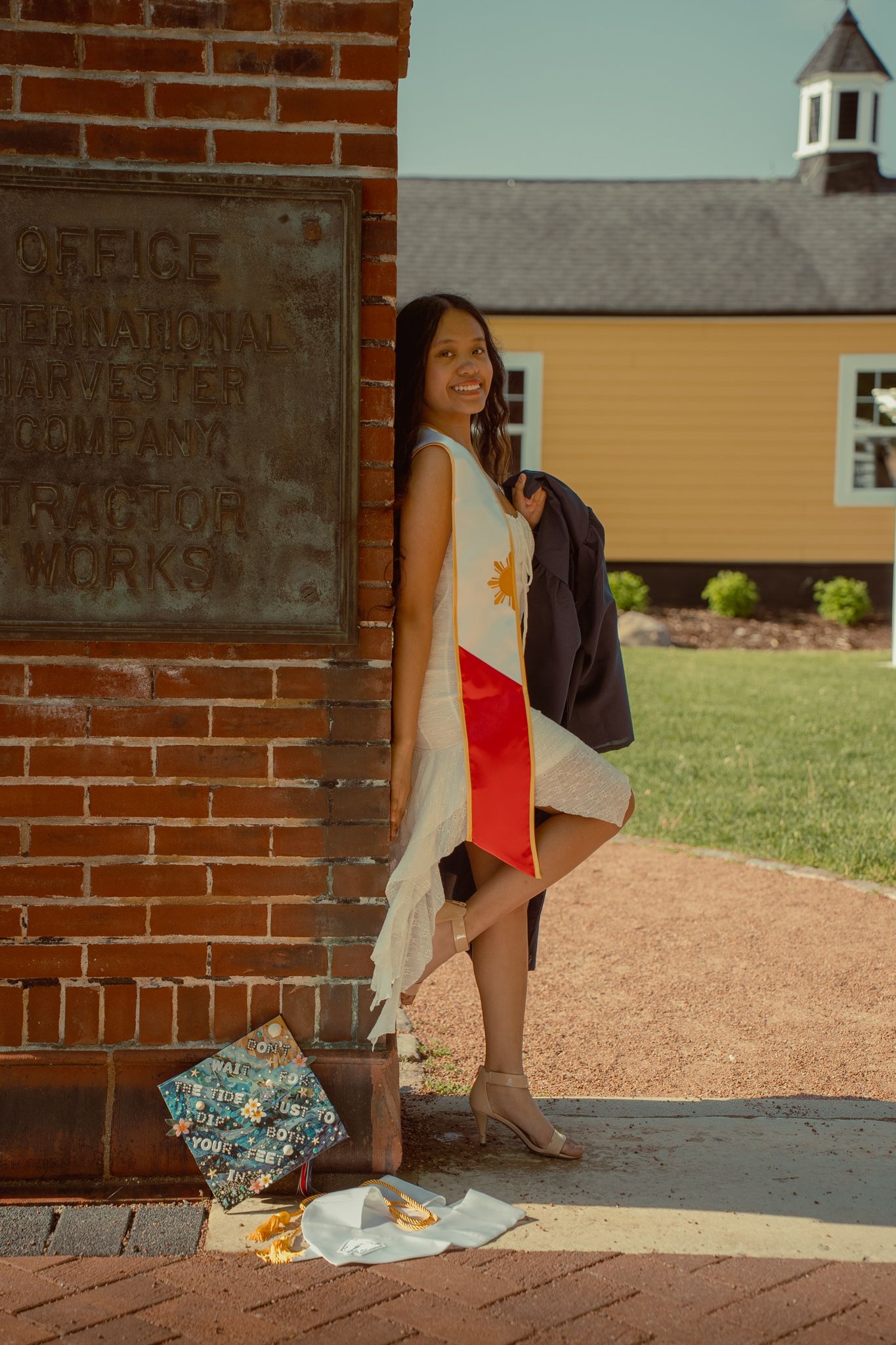 Smiling graduate wrapped in Philippine flag holding academic gown beside brick building on sunny day.
