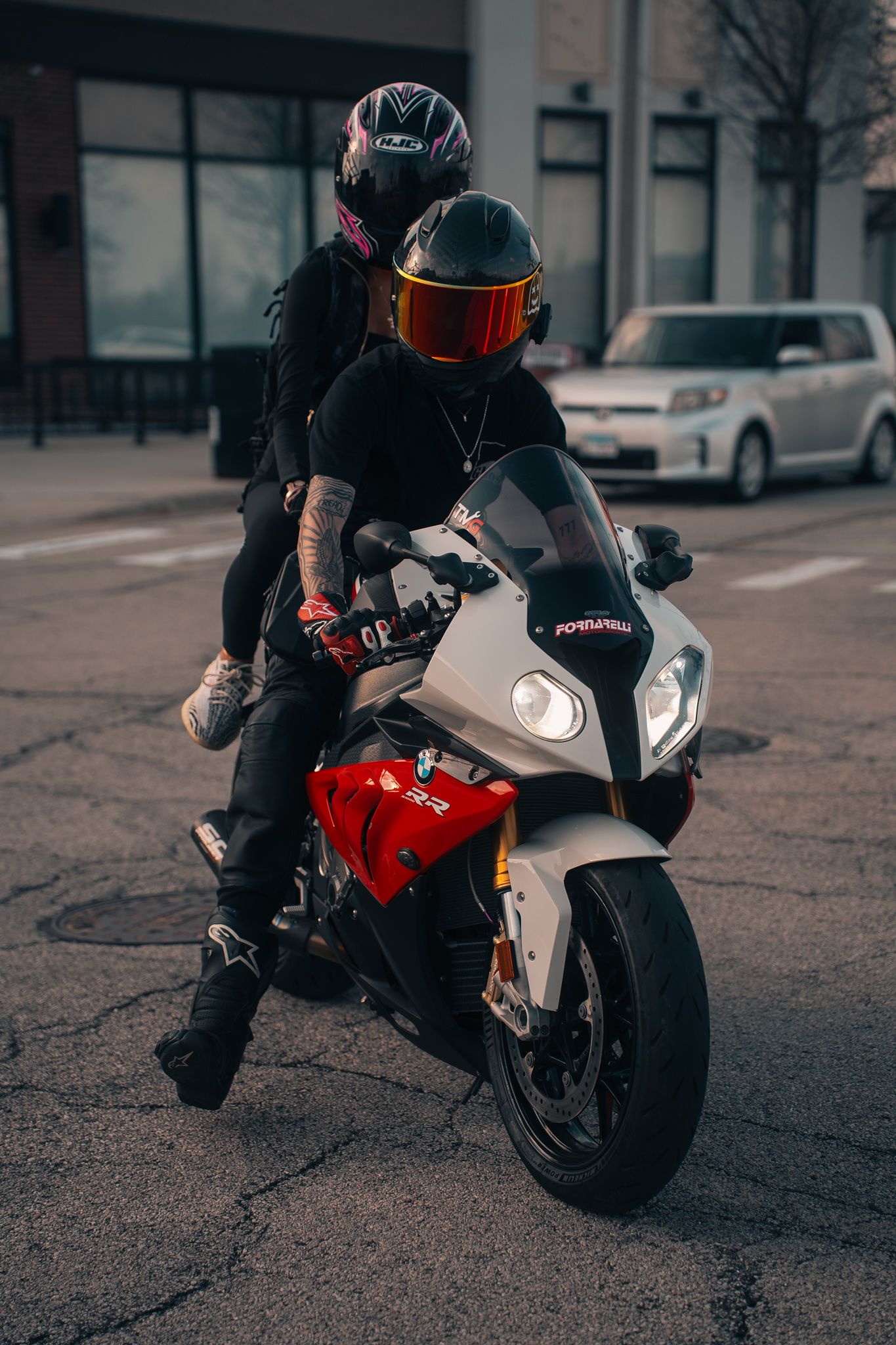 Two riders in helmets pose on a red and white BMW S1000RR sport motorcycle on an urban street.