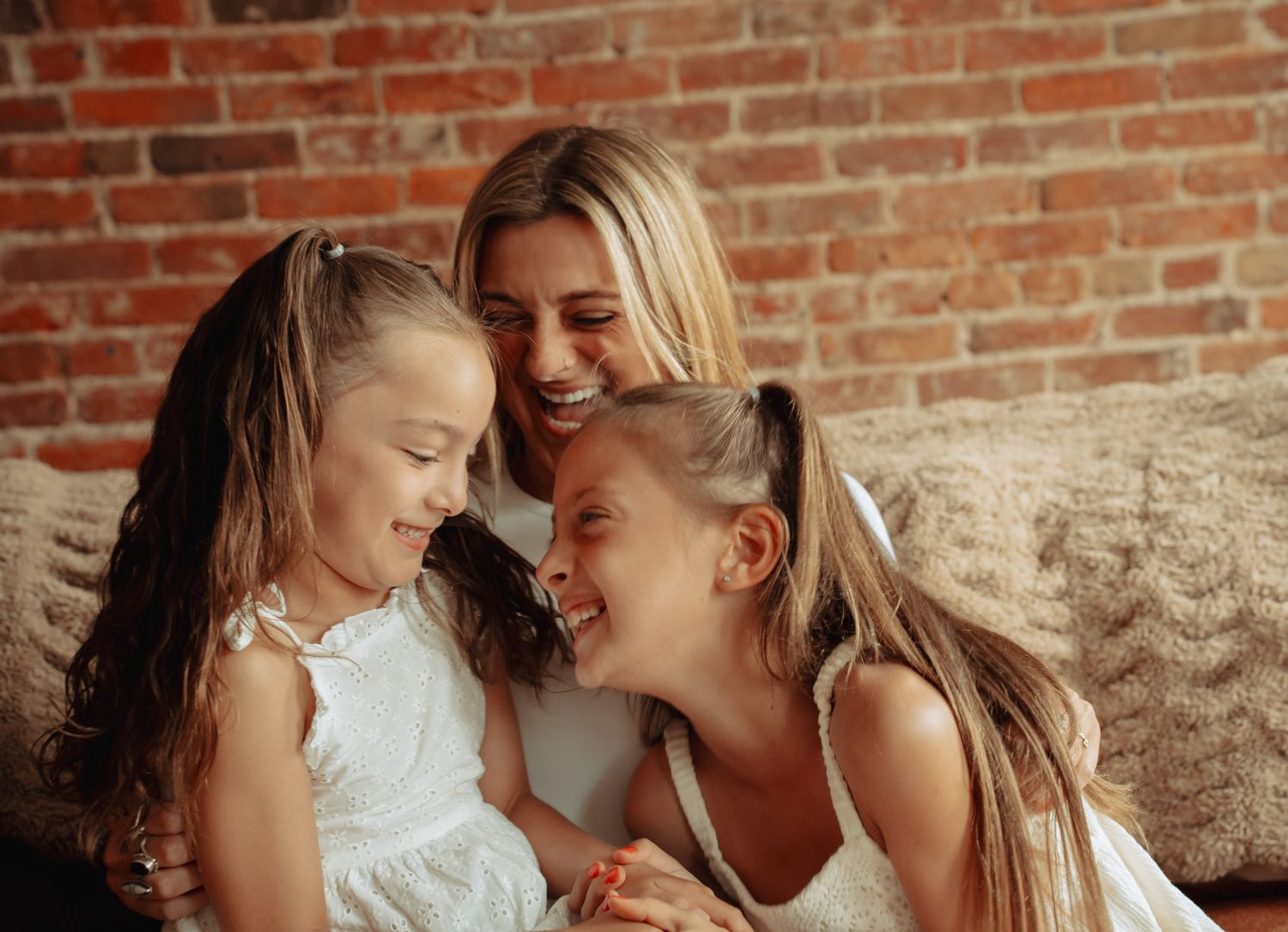 A mother laughs joyfully with her two young daughters in white dresses, sharing a warm embrace on a couch.