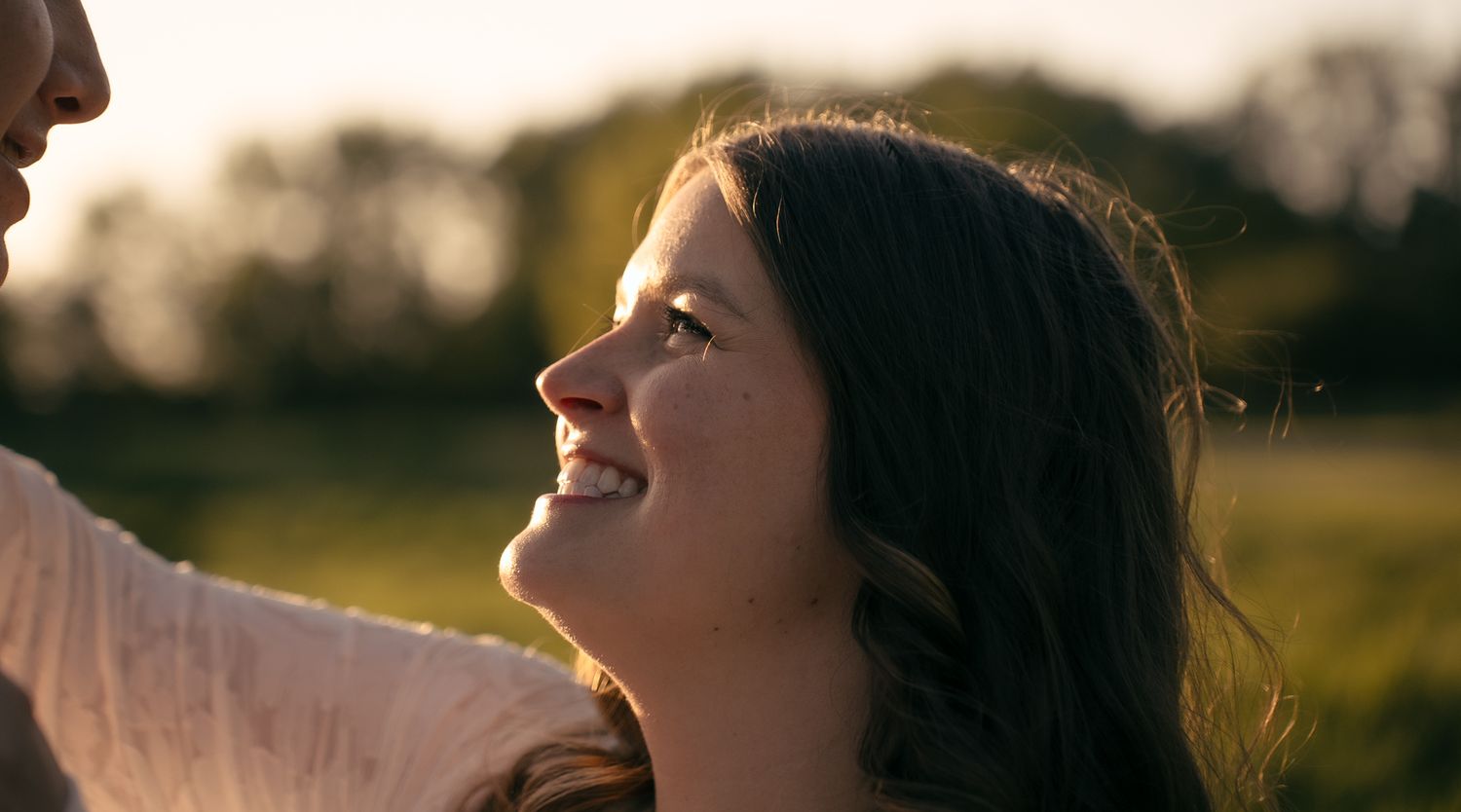 Young woman smiling warmly in golden sunlight outdoors, with lush green trees in the background.