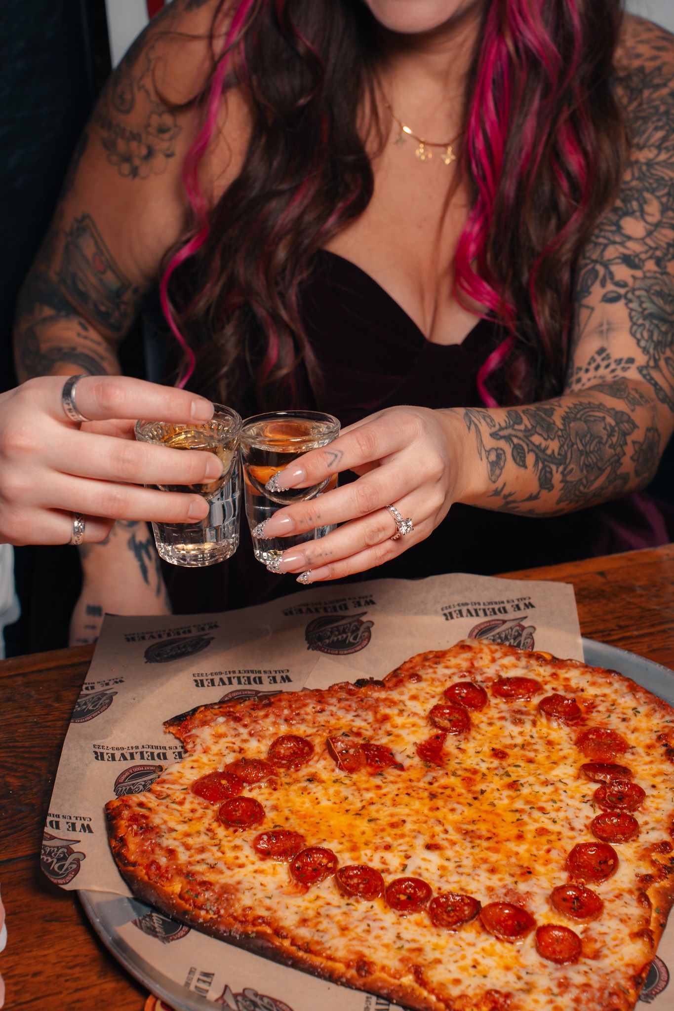 Heart-shaped pepperoni pizza being shared with tattooed hands reaching for slices on a rustic wooden table.