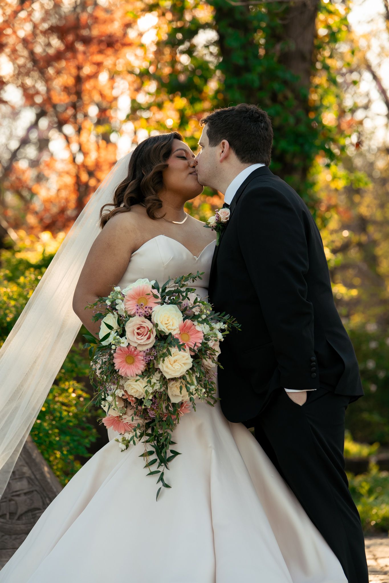 Bride and groom share a kiss outdoors, bride holding a cascading pink and white bouquet, surrounded by autumn foliage.