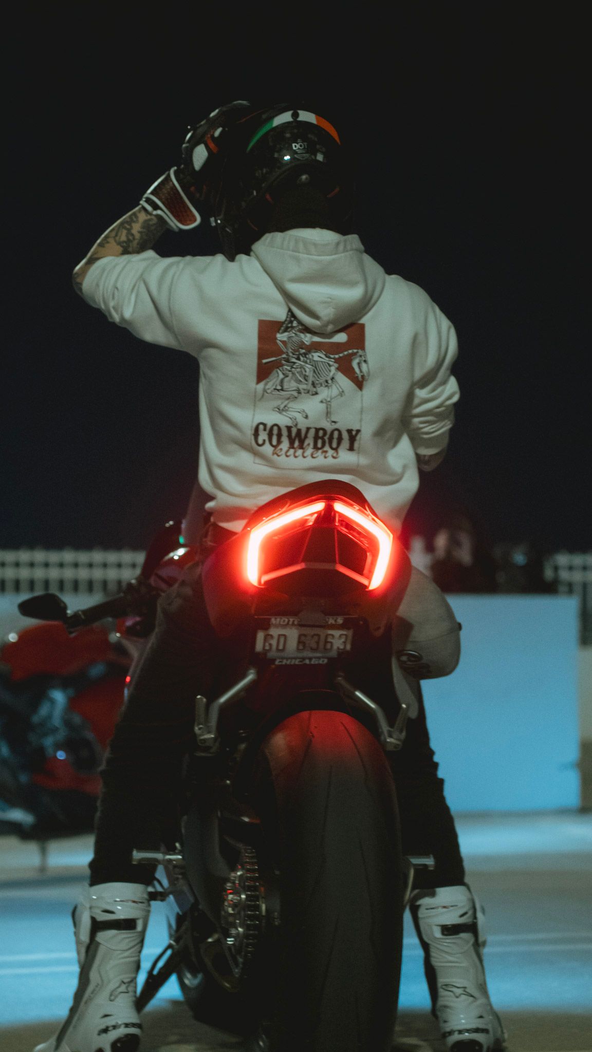 Motorcycle rider wearing white jacket with red taillights glowing in dark setting.