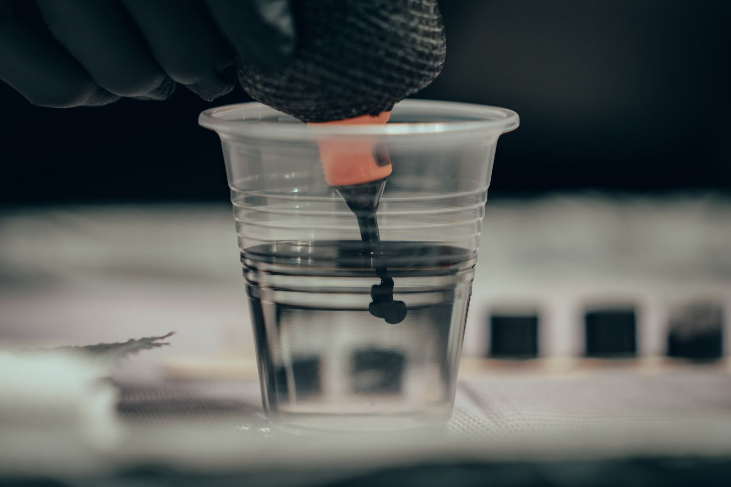 Gloved hand dripping black ink into a clear plastic cup, close-up shot in a tattoo studio setting.