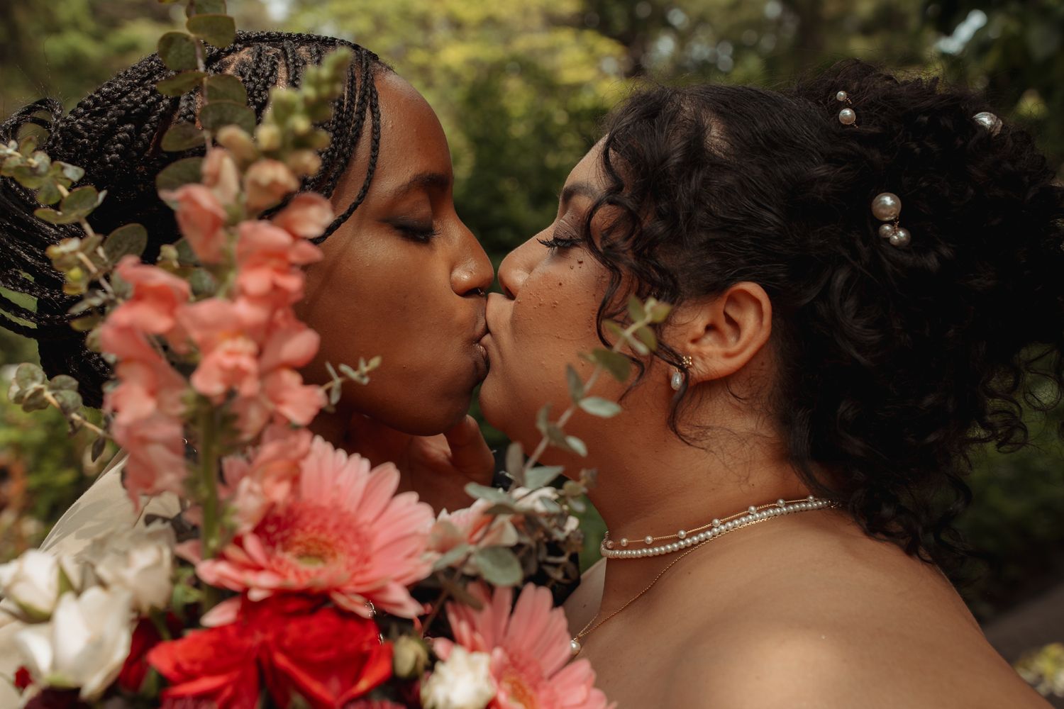 Two brides sharing a kiss surrounded by a vibrant bouquet of pink and red flowers at their wedding.