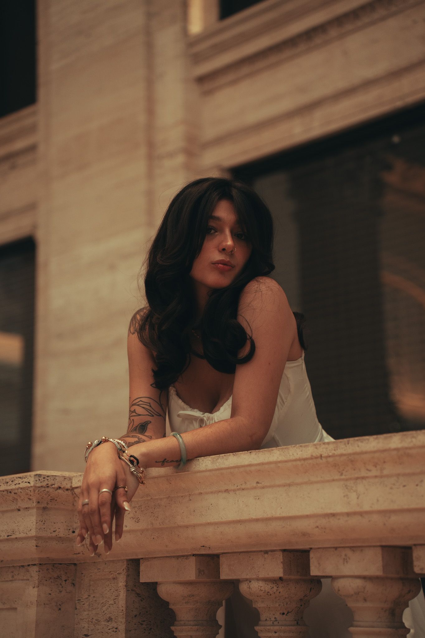 A woman in a white top leans on a stone balcony railing at dusk, wearing bracelets and showing tattoos.