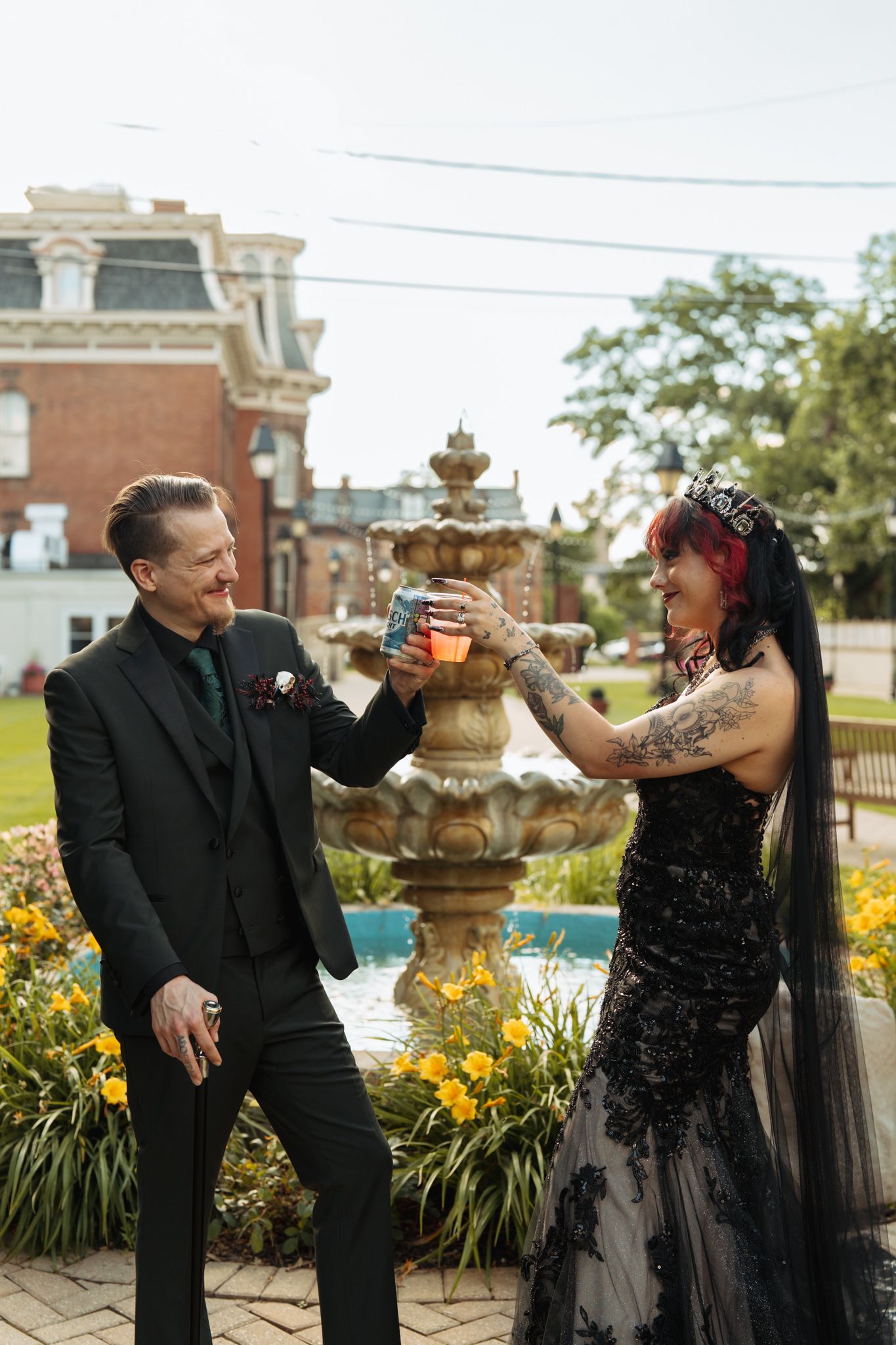 Gothic bride in black lace gown and groom in black suit toasting drinks by ornate fountain at outdoor wedding.