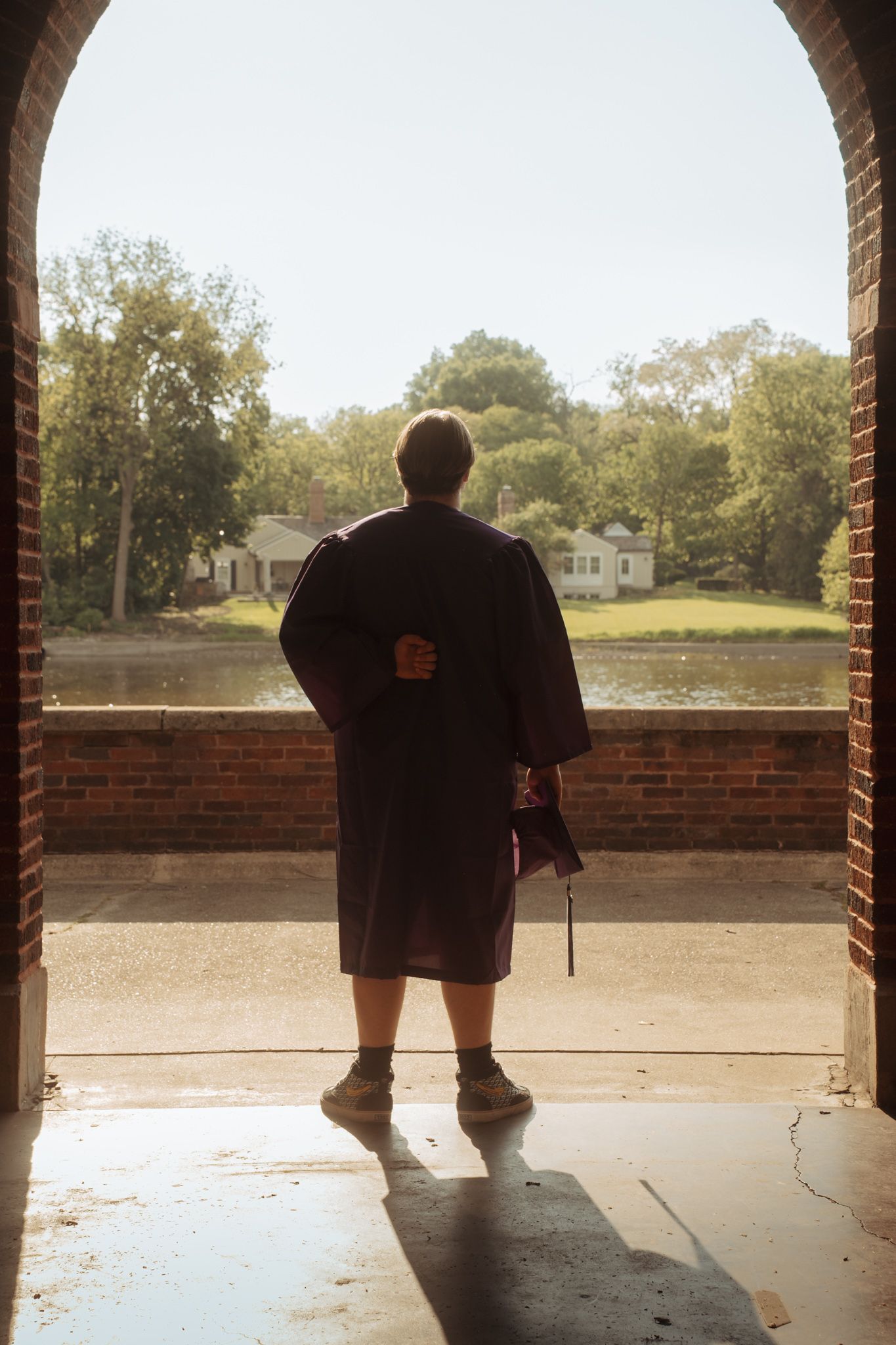 Graduate in robe stands under brick arch overlooking serene lake and trees, holding diploma after commencement ceremony.