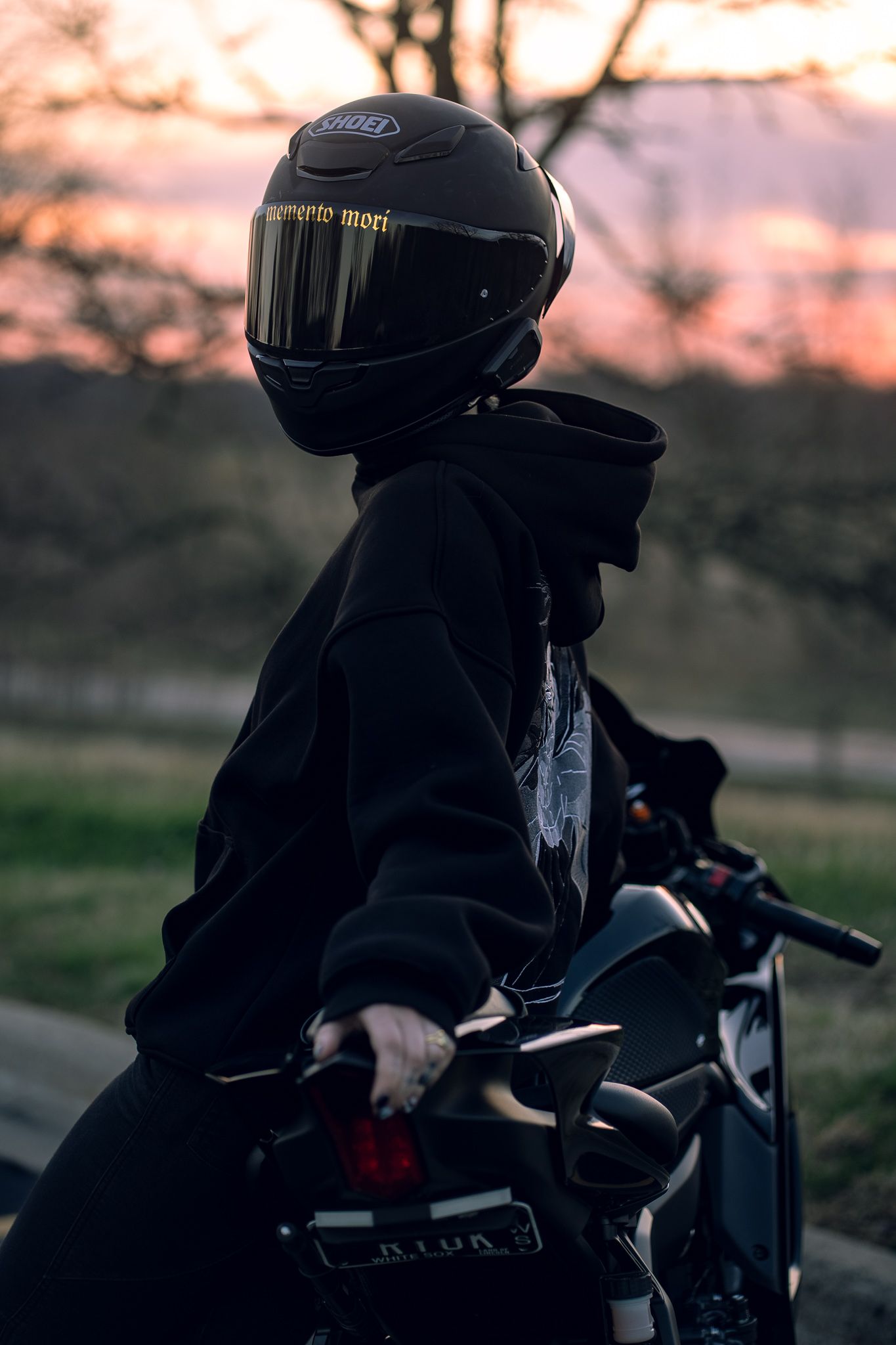 Motorcyclist in black hoodie and Shoei helmet sits on bike at sunset with golden sky backdrop.