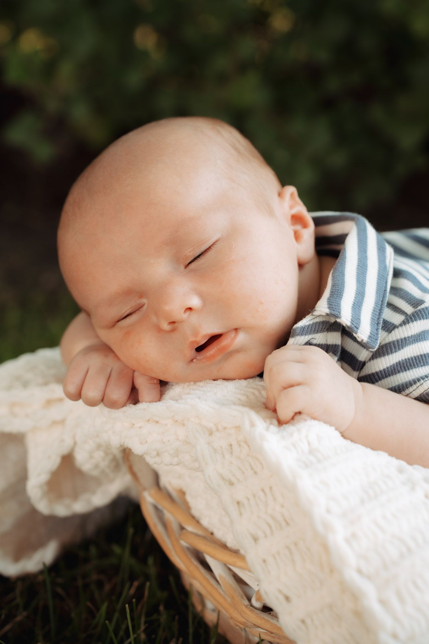Peaceful newborn baby sleeping in a wicker basket outdoors, wearing a blue striped onesie with a soft white blanket.