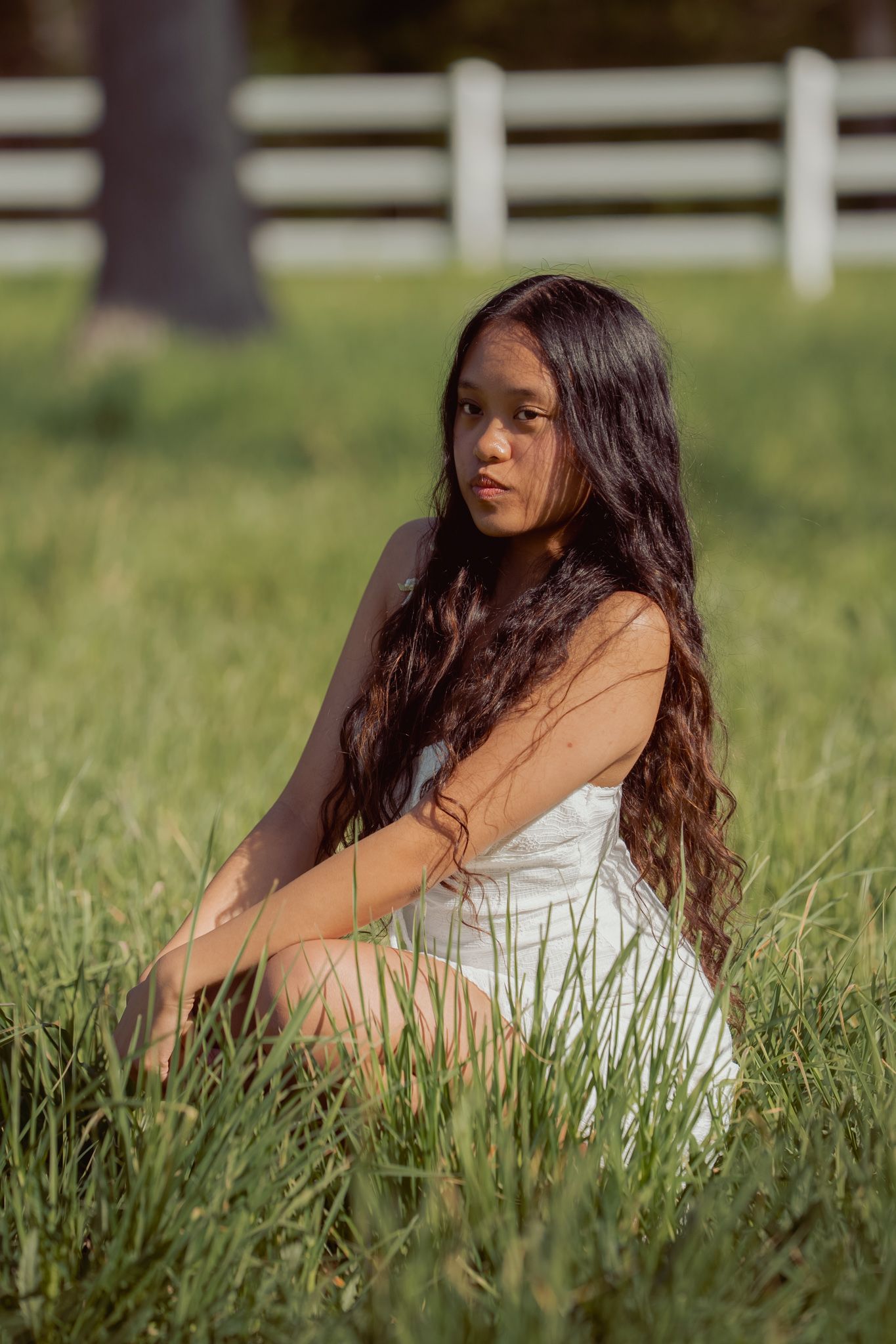 Young woman with long wavy hair sits in green grass wearing a white dress, with a white fence and trees in the background.