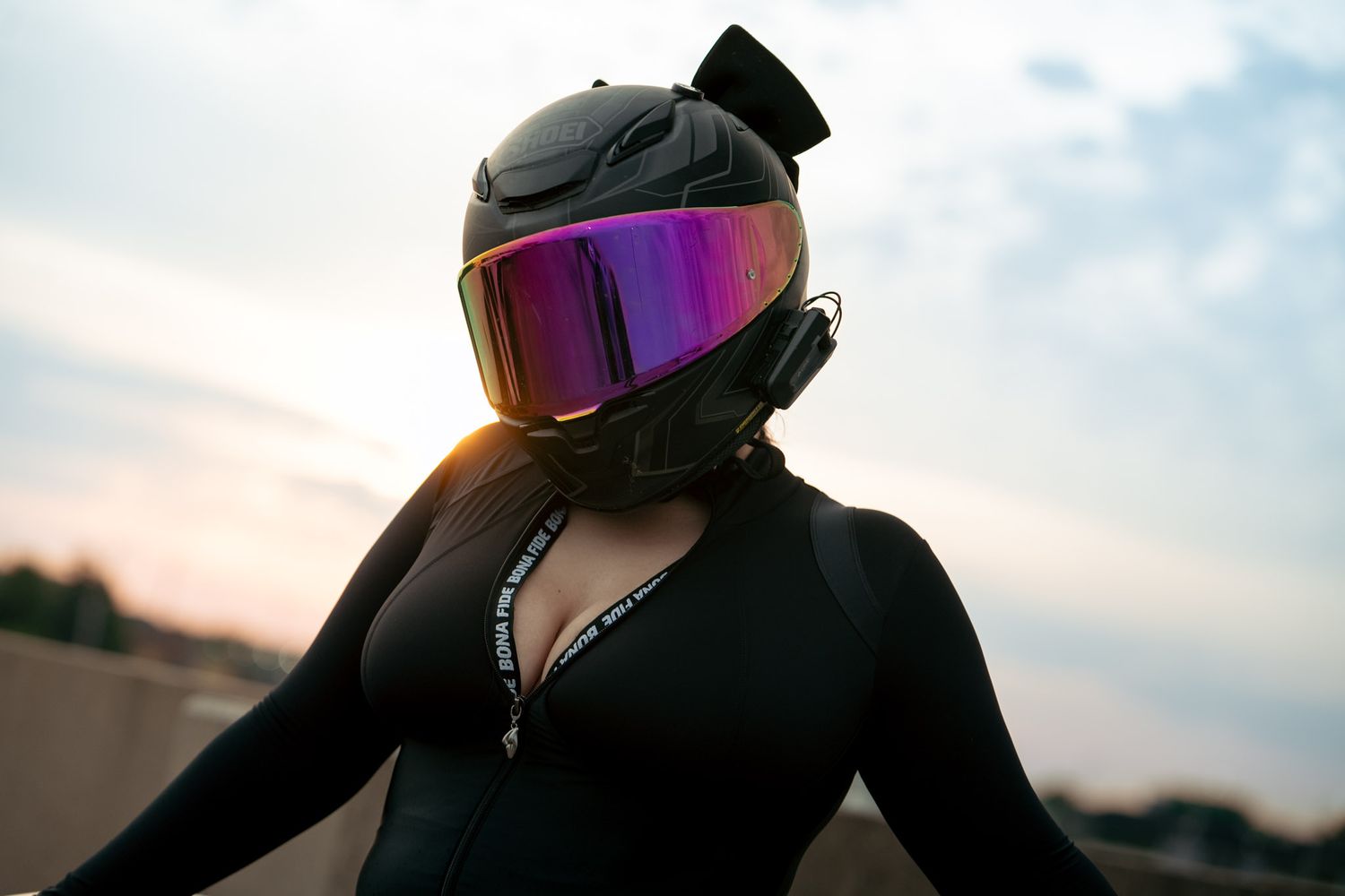 Person wearing a black motorcycle helmet with iridescent visor and black bodysuit posing outdoors at sunset.