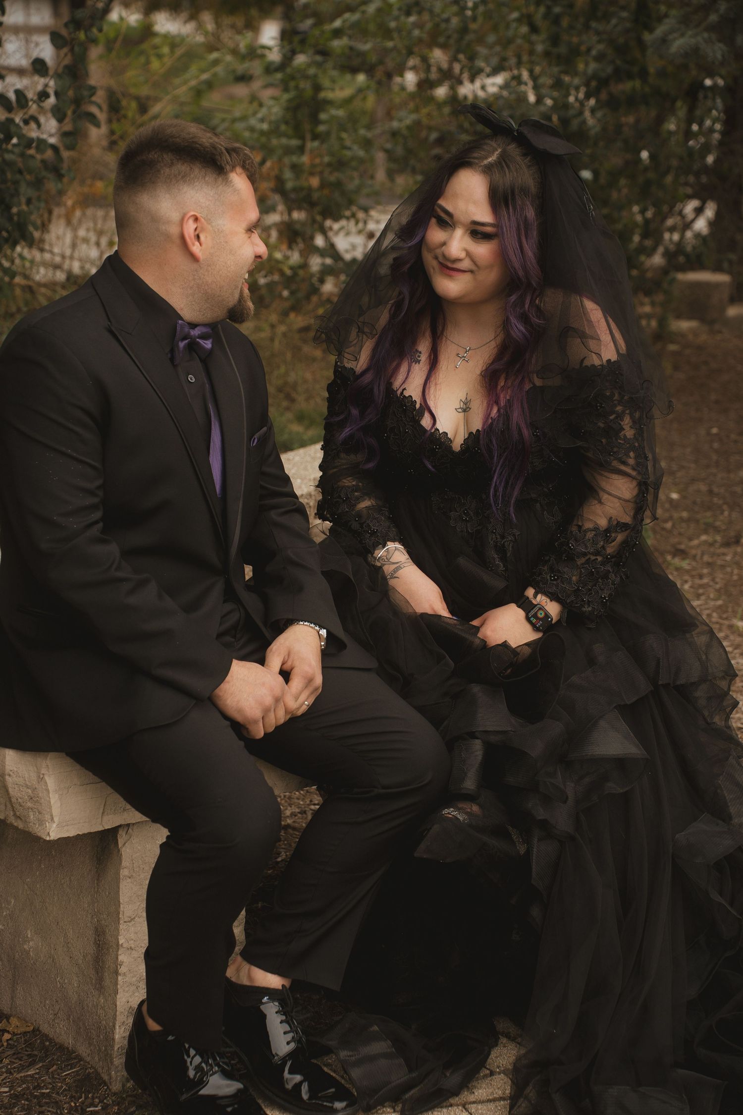 Couple in gothic attire sits intimately on stone bench in autumn setting.